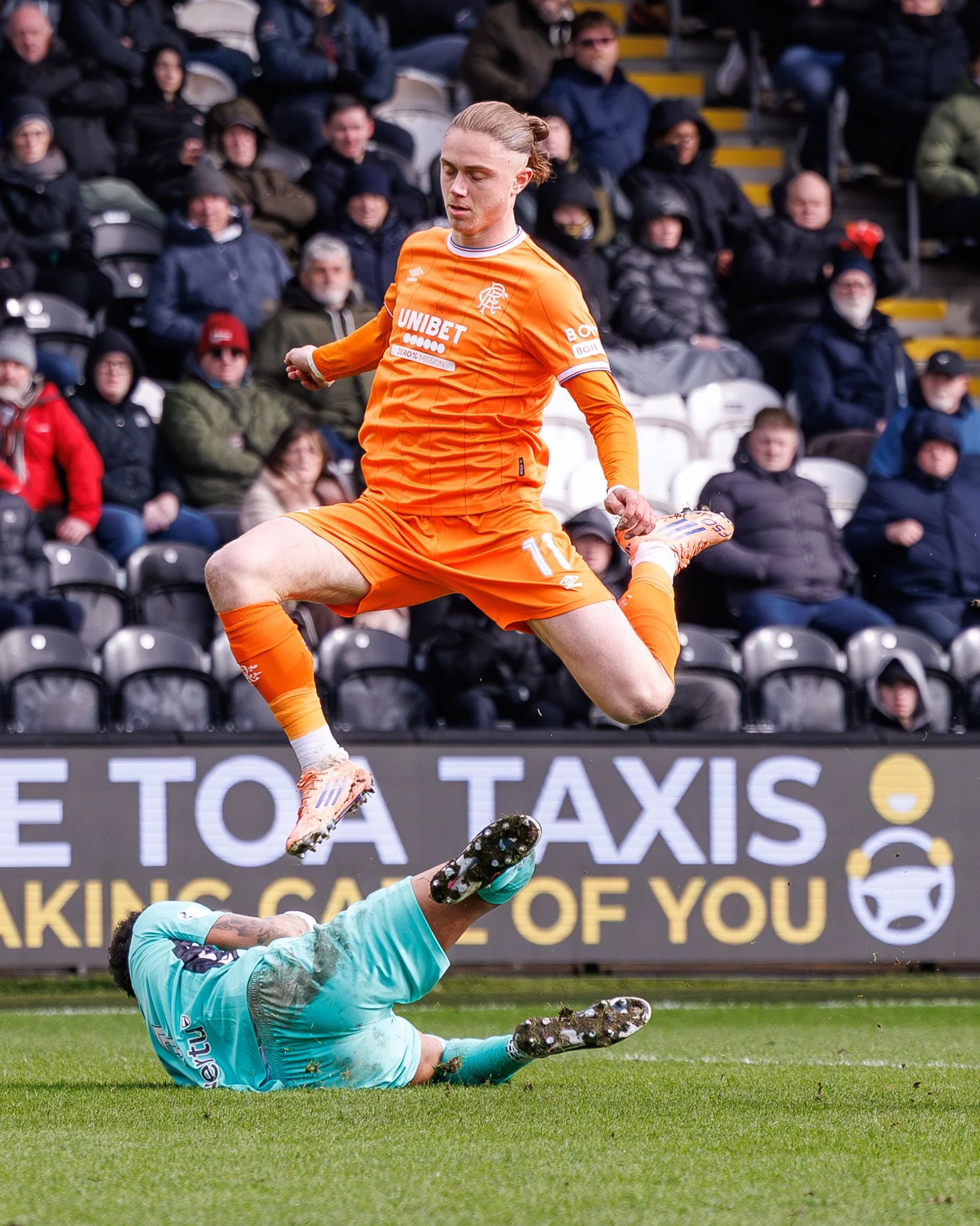 A soccer player in an orange uniform is kicking a soccer ball, while a goalkeeper in a teal uniform is on the ground after attempting to save the shot, with spectators watching in the background.