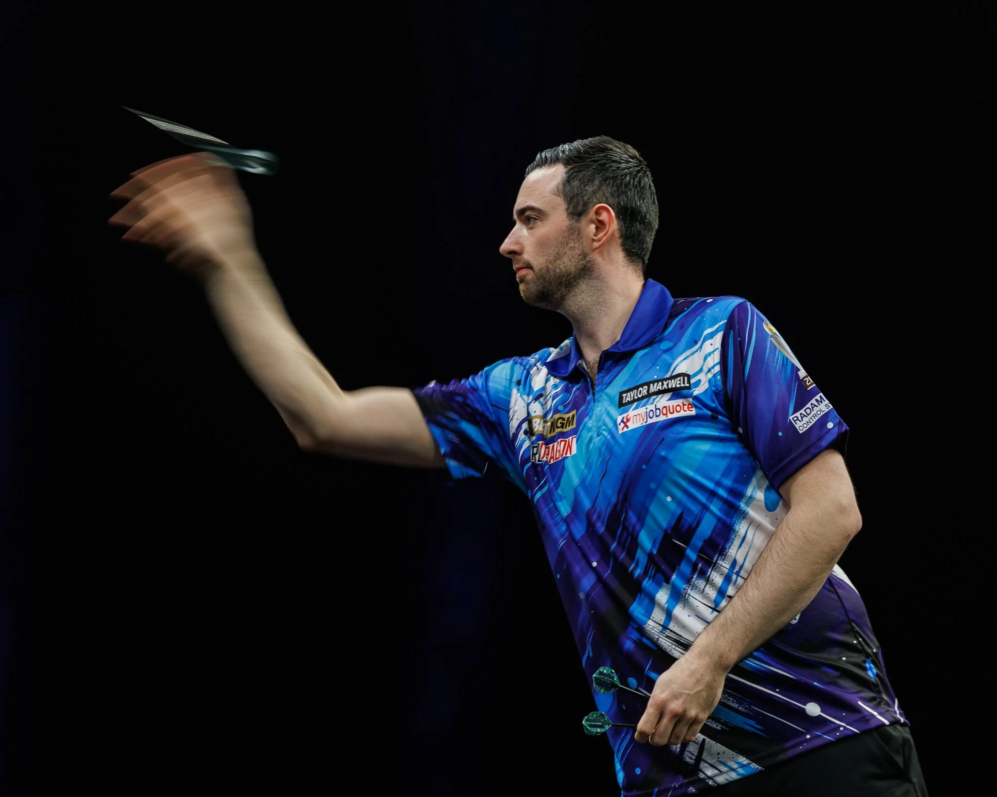 A man with dark hair, wearing a blue sports jersey with various sponsorship logos, is holding a dart and appears to be in the process of throwing it in a dark setting.