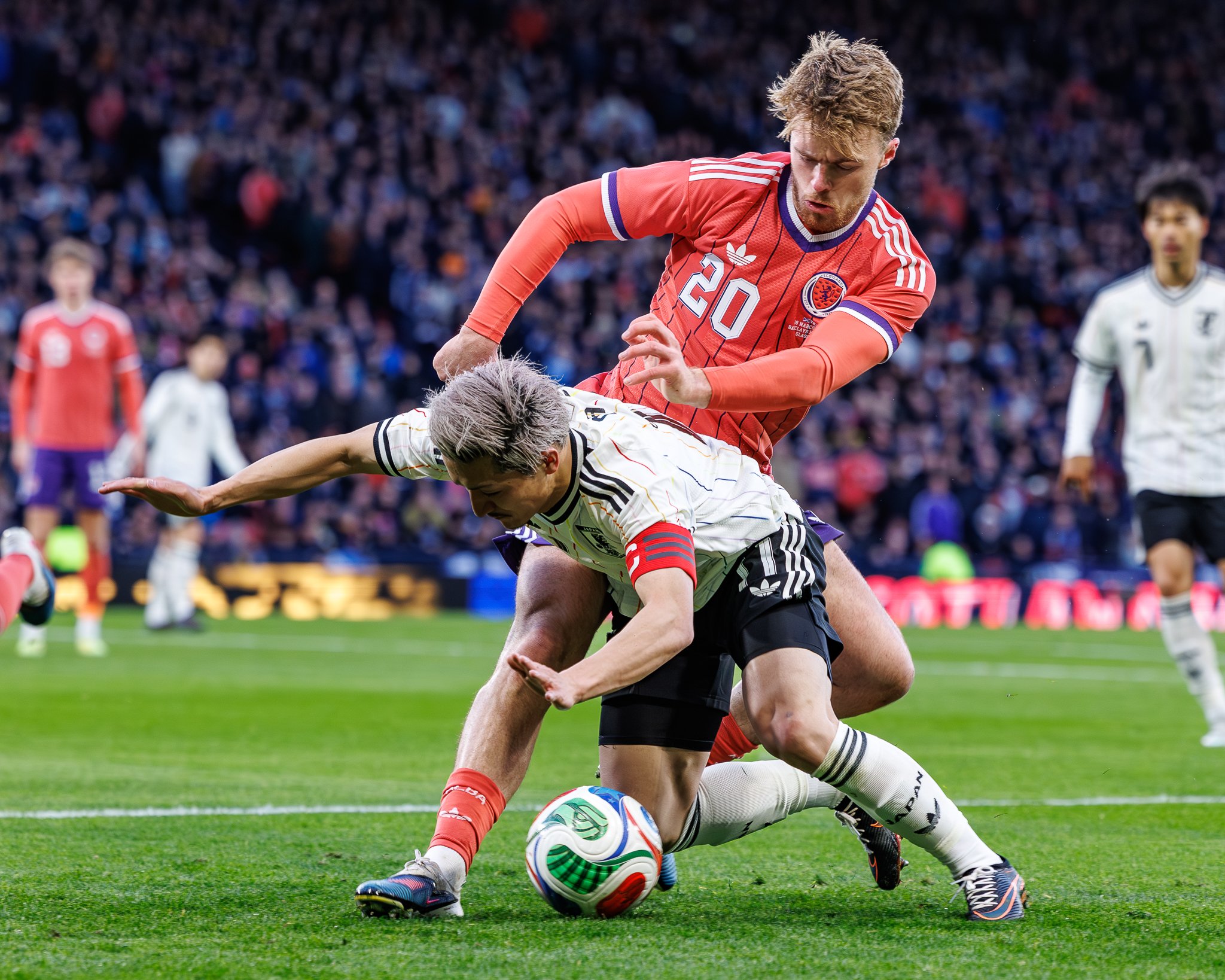 A soccer player in a red jersey with the number 20 is tackling a player in a white jersey with black stripes during a match, with a crowd in the background.