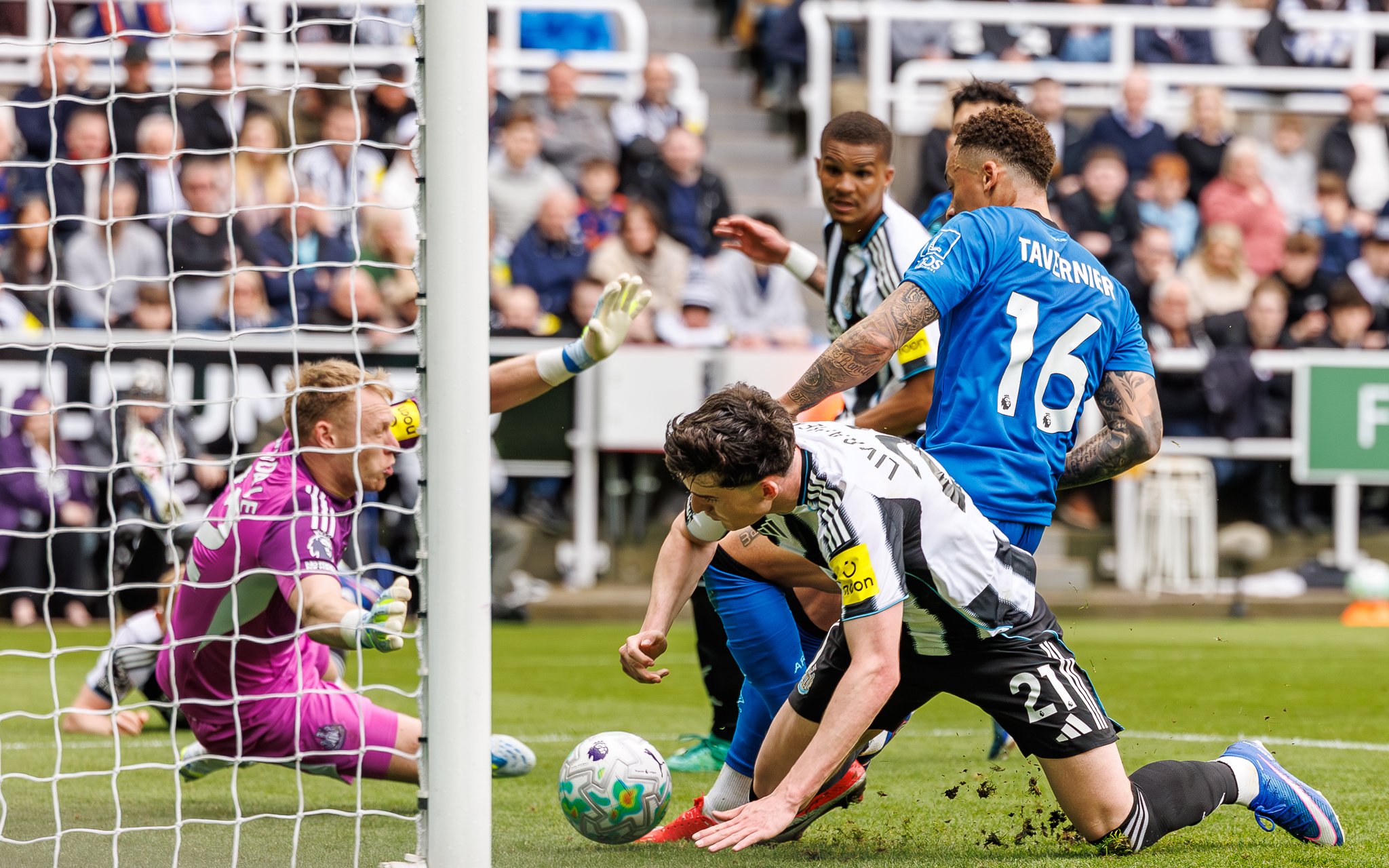 Soccer players competing for the ball near goal, with goalkeeper in pink covering the goal, during a match with spectators in the background.