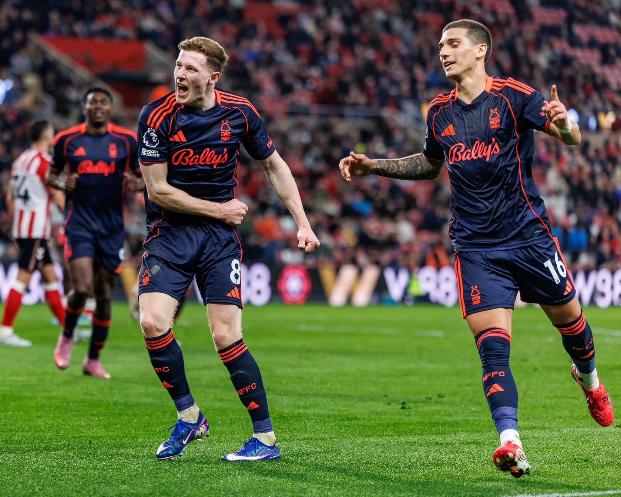 Soccer players in blue and red uniforms celebrate on the field during a match, with a crowd of spectators in the background.
