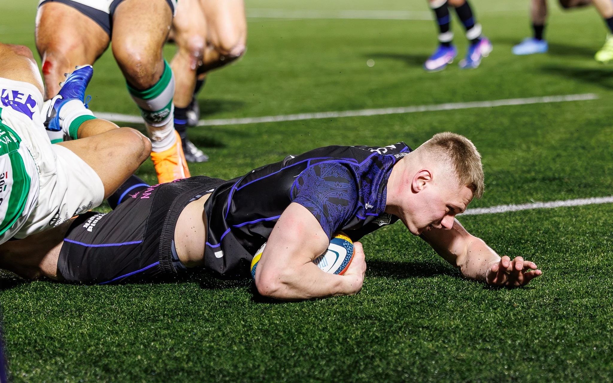 A rugby player in black and purple uniform is lying on the field holding a rugby ball, while a player in white and green is on top, trying to tackle him during a game.