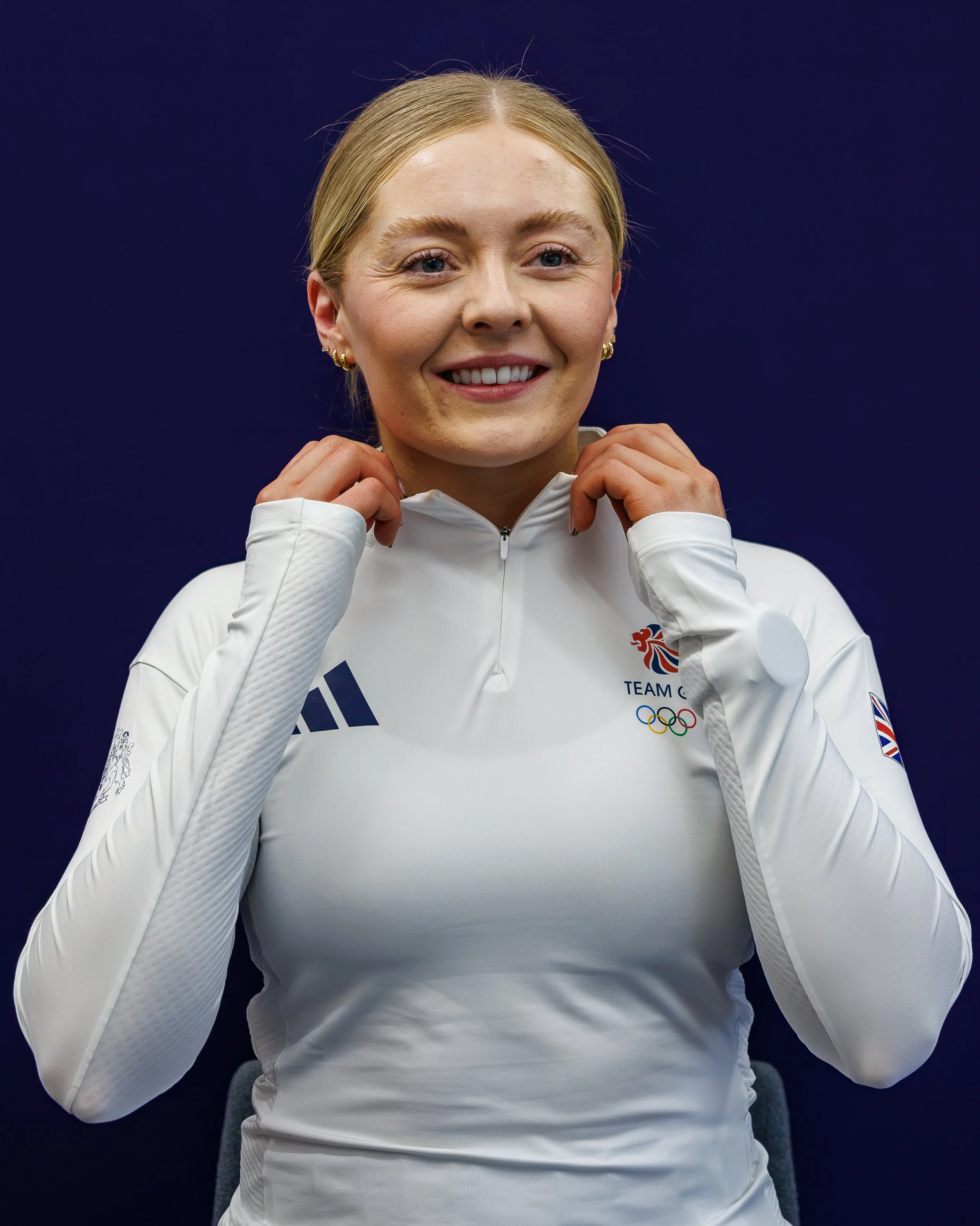 A smiling woman in a white sports jacket with Team GB and Olympic logos, sitting against a dark background.