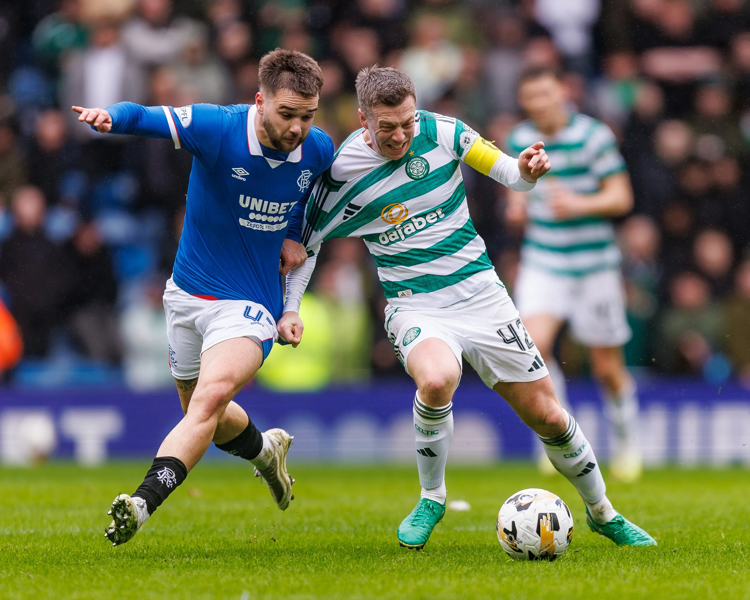 Two soccer players competing for the ball during a match, one in a blue jersey and the other in a green and white striped jersey, on a grassy field with blurred spectators in the background.