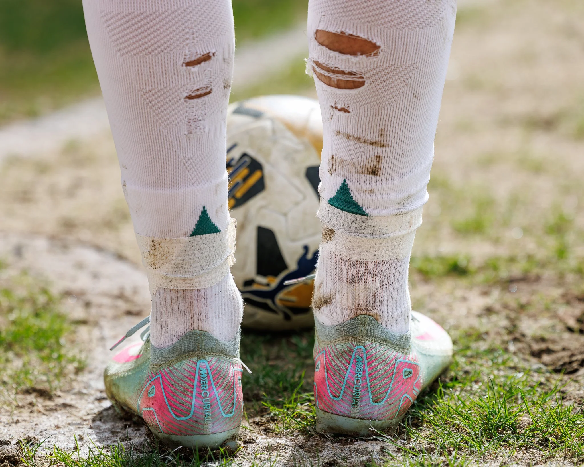 Close-up of two children's legs wearing torn white leggings with trees printed at the bottom, pink and blue sneakers, and muddy soles. A soccer ball is partially visible on the ground between their legs on a grassy and dirt outdoor field.