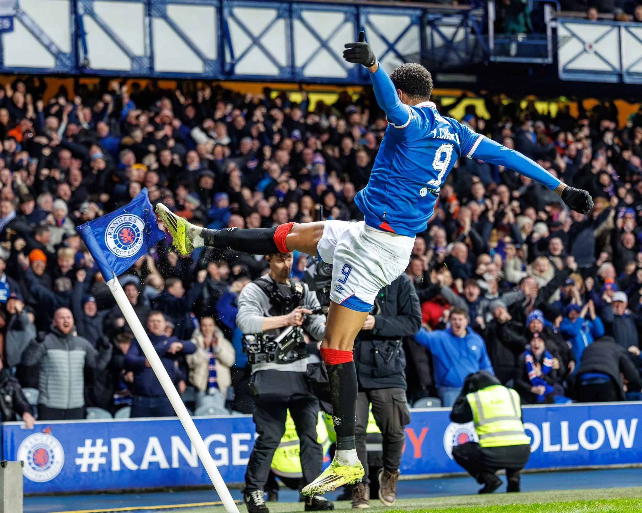 A soccer player in a blue uniform celebrates by jumping with arms outstretched near the corner flag, with a large crowd of spectators in the background.