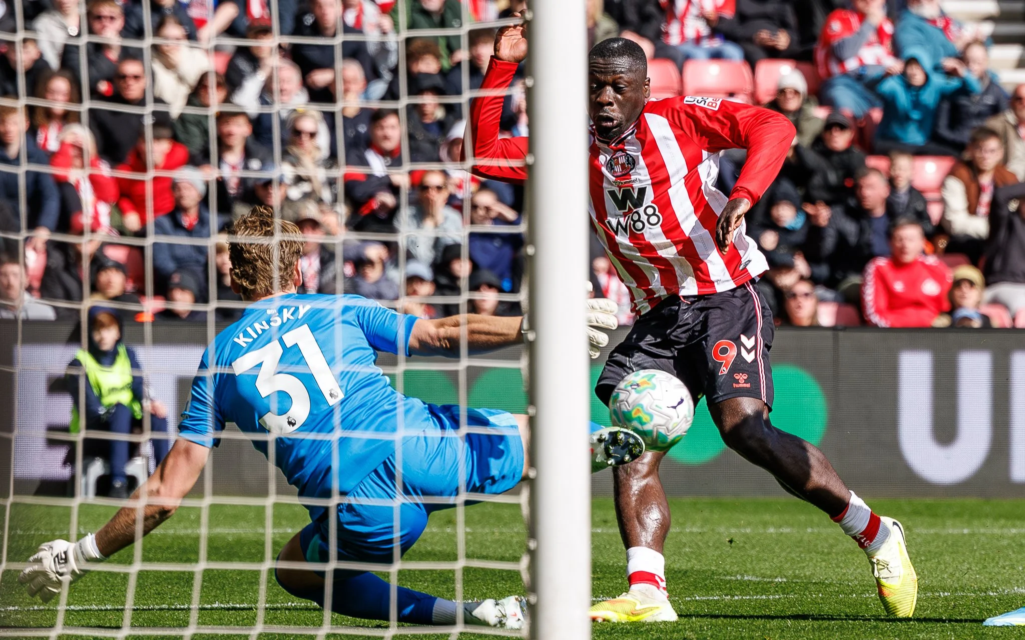 A soccer player in a red and white striped jersey dribbling the ball near the goal, with a goalkeeper in a blue jersey making a save attempt during a match attended by a crowd.