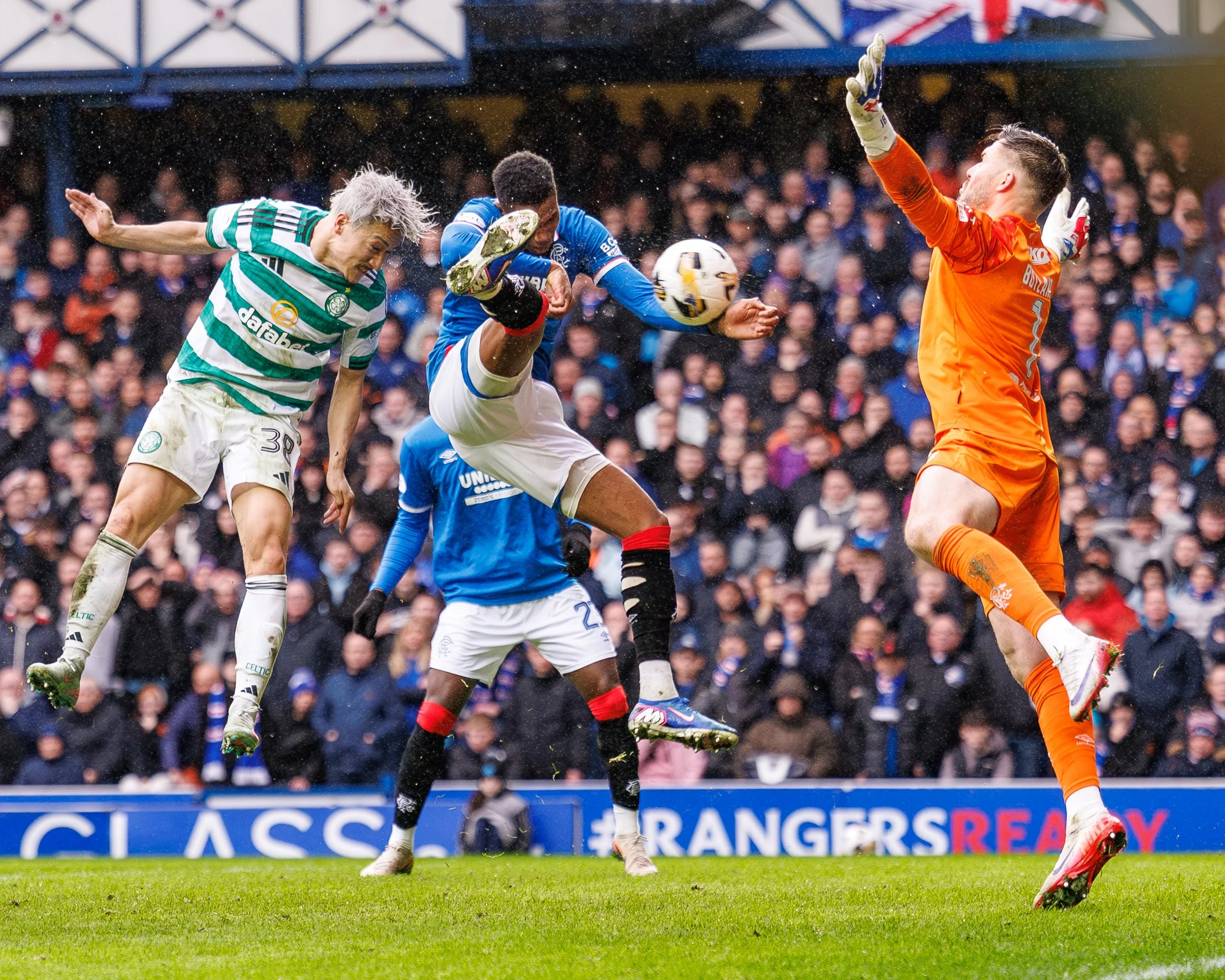 Soccer players in action during a match, with a goalkeeper in orange attempting to block a shot, and two outfield players jumping to head the ball amidst rain, crowd in the background.