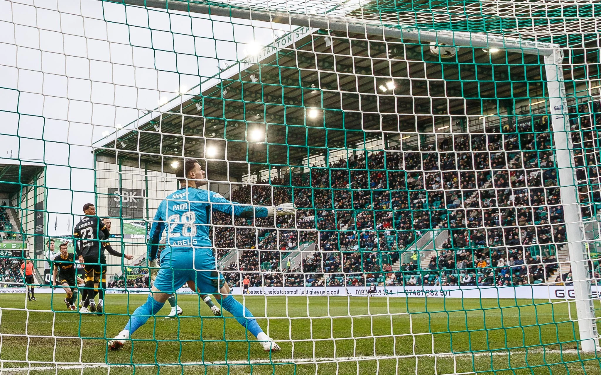 A soccer game taking place at a stadium with a goalkeeper in a blue uniform and two players from opposing teams near the goal, with a large crowd in the background.