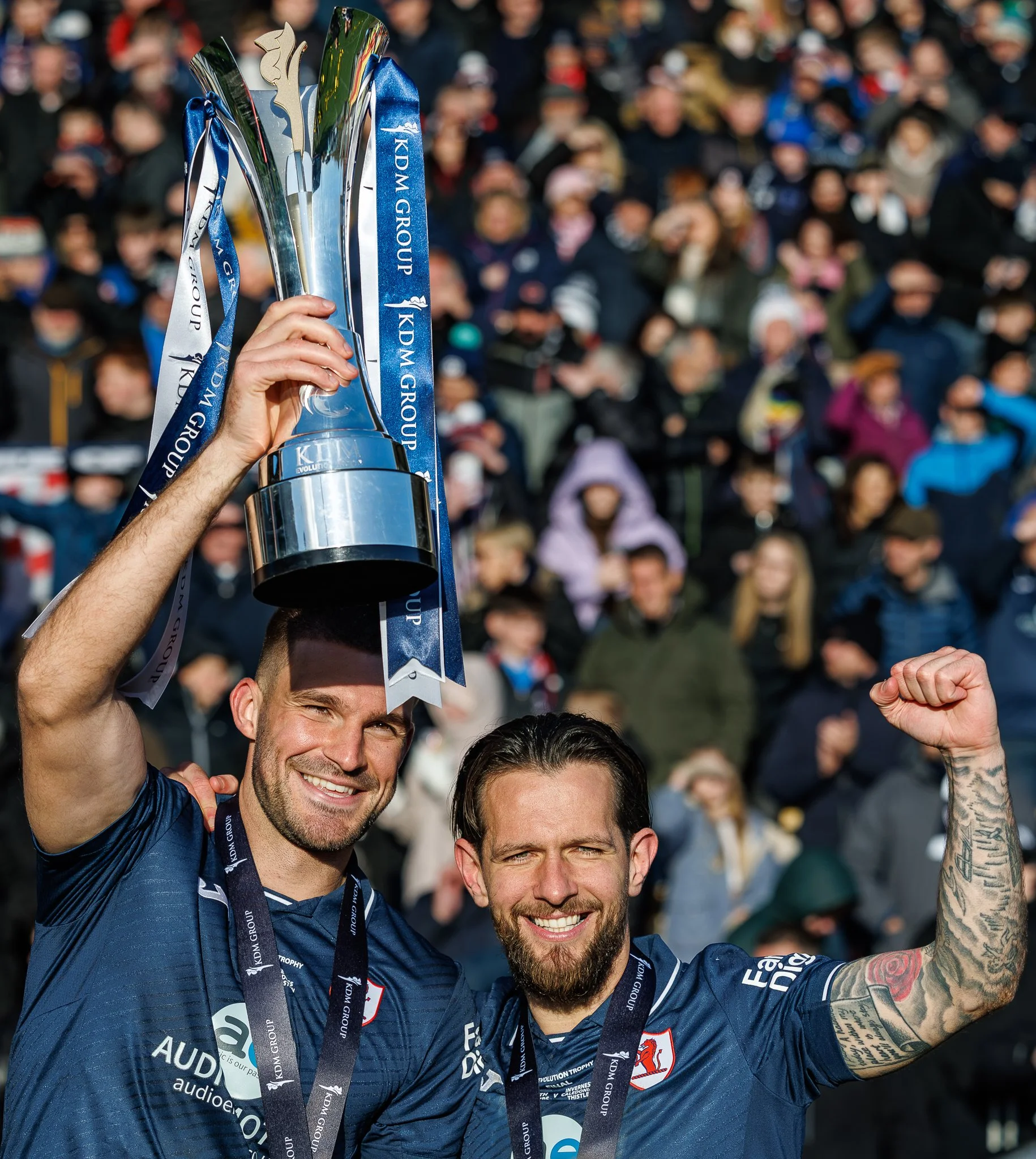 Group of men celebrating with trophies at an outdoor event, one holding up a trophy while smiling and making a fist gesture, with a crowd in the background.