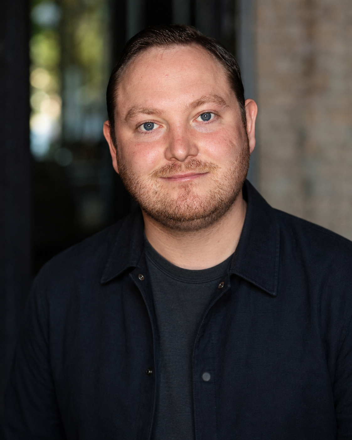 Close-up portrait of a young man with light skin, short brown hair, blue eyes, and a beard, wearing a black shirt and smiling at the camera. Blurred indoor background with natural light.
