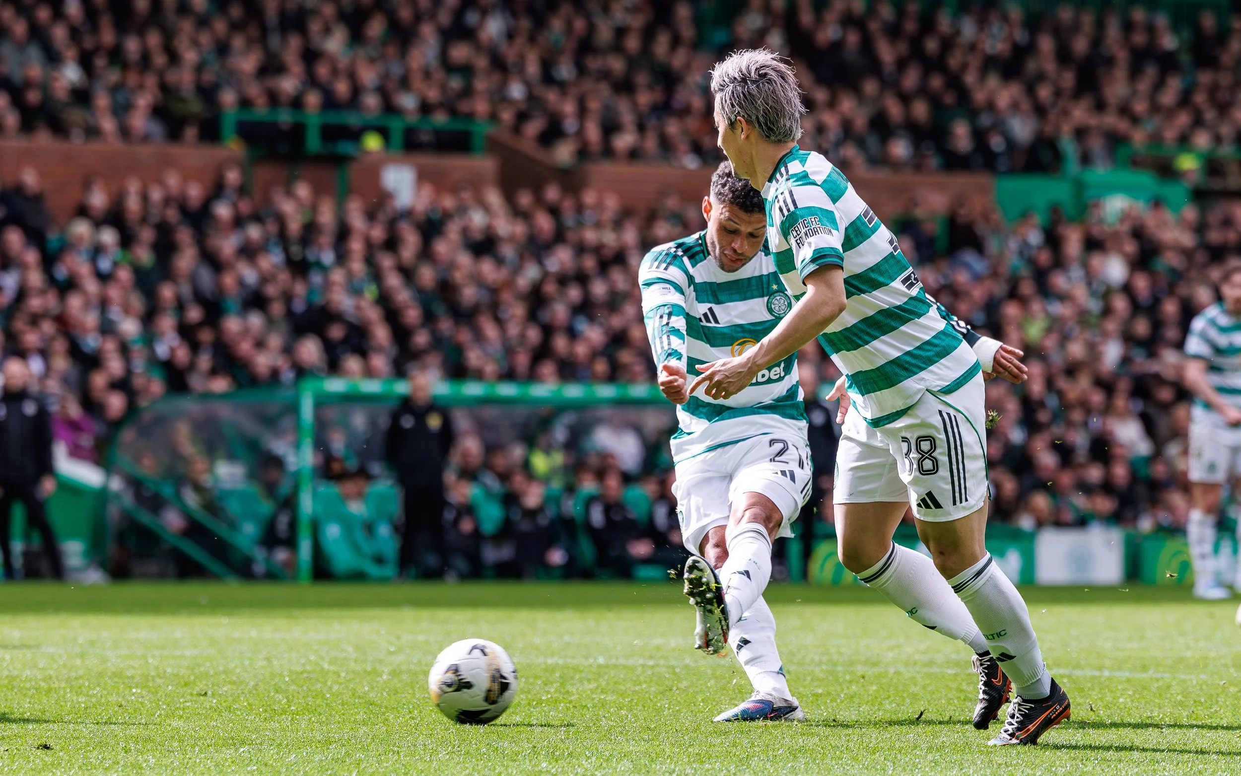 Two soccer players from Celtic FC, one with gray hair, are on a green field during a match, facing a large crowd in the stadium.