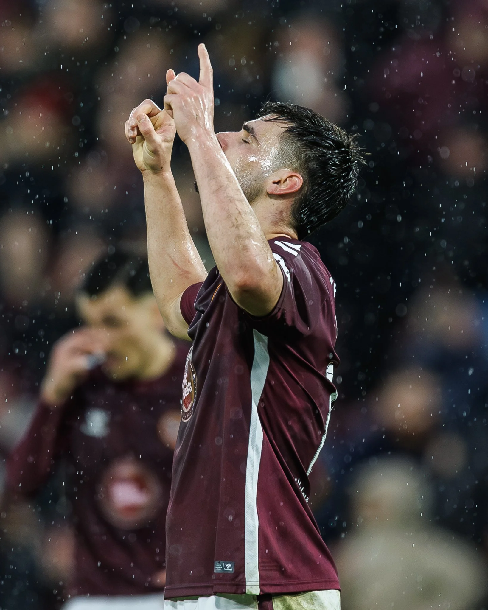 A soccer player celebrating on the field during a match, wearing a maroon and white uniform, with water droplets on his face and hands.