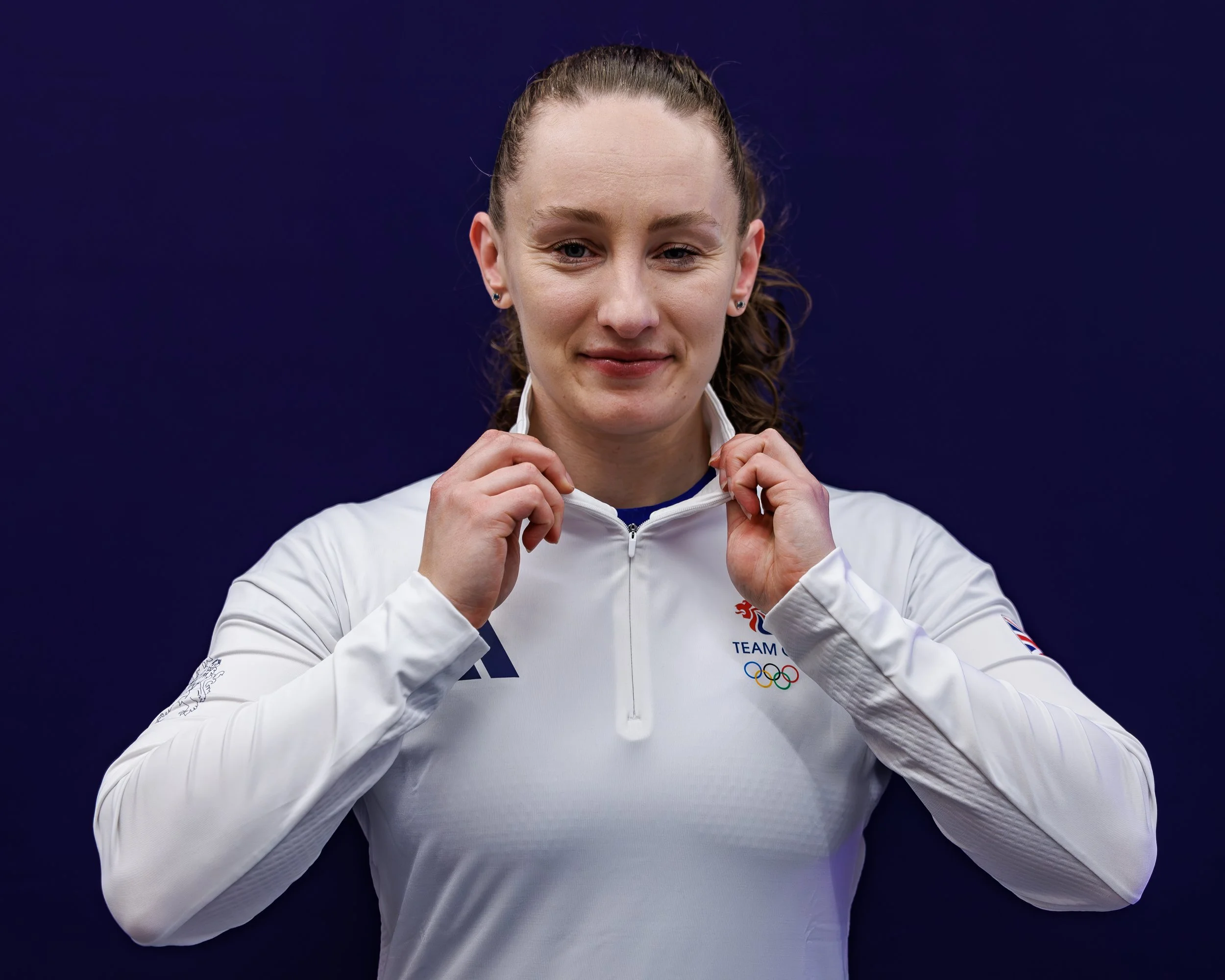 A female athlete with red hair pulled back, wearing a white jacket with the British Olympic logo, adjusting her collar, against a dark background.