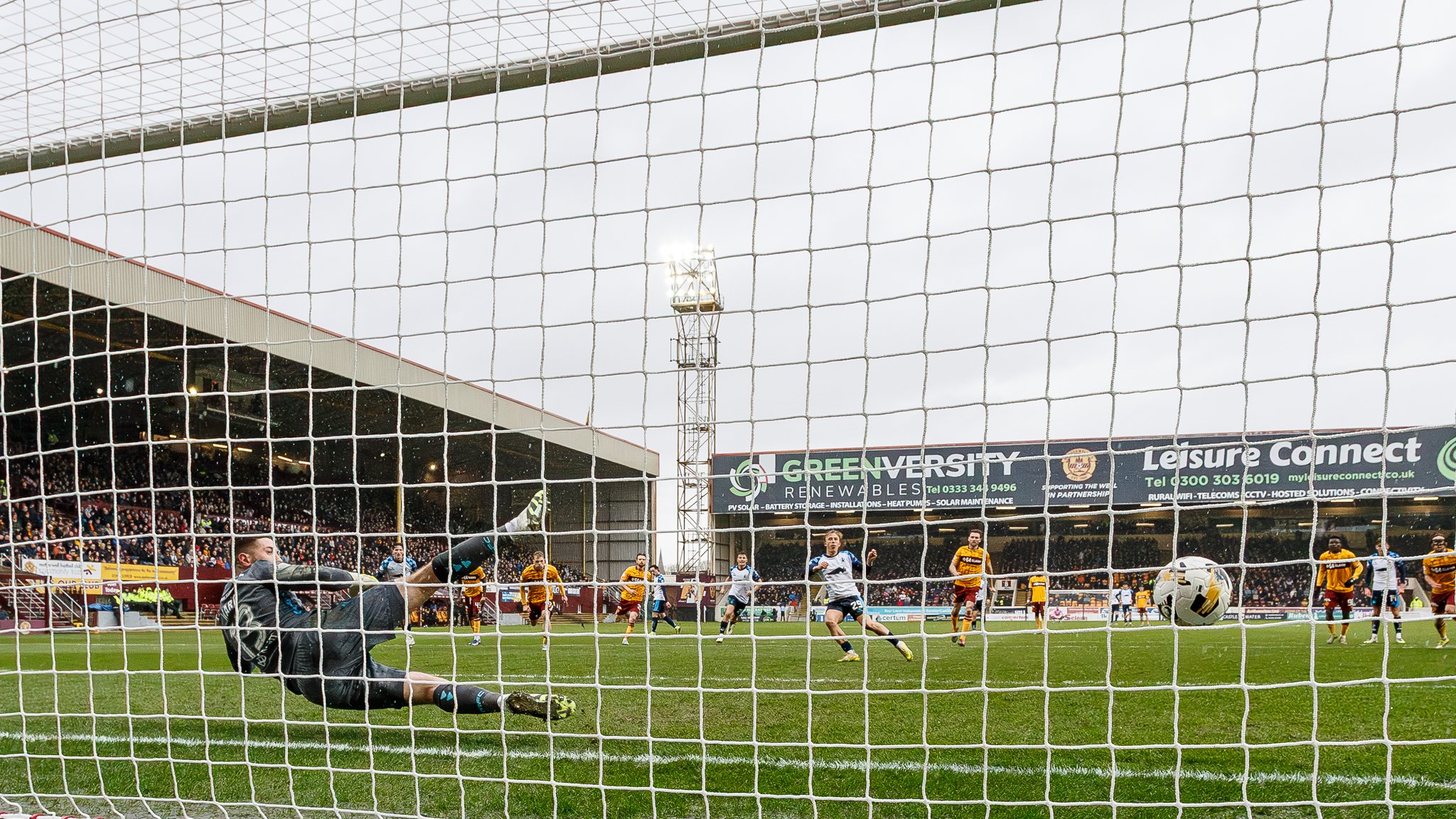 A goalkeeper diving to make a save during a soccer match viewed through a goal net. Players and spectators are visible in the background.