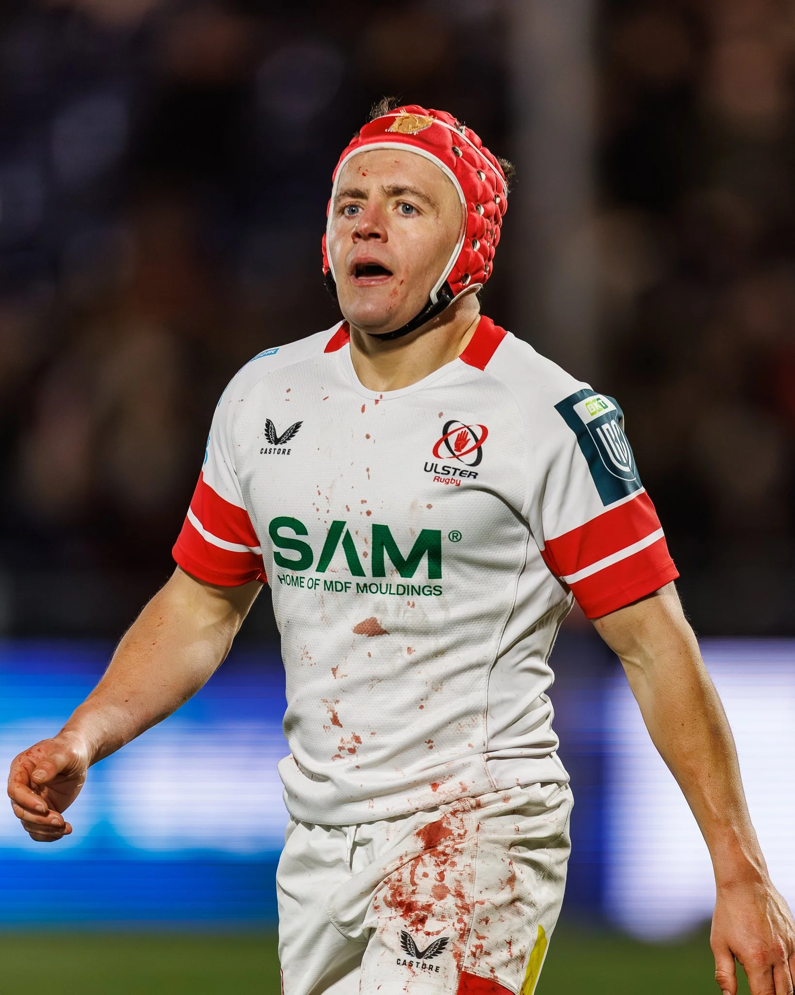 A rugby player wearing a white uniform with red accents and bloodstains on the shirt, a red protective headgear, with an expression of surprise or fatigue, on a rugby field.