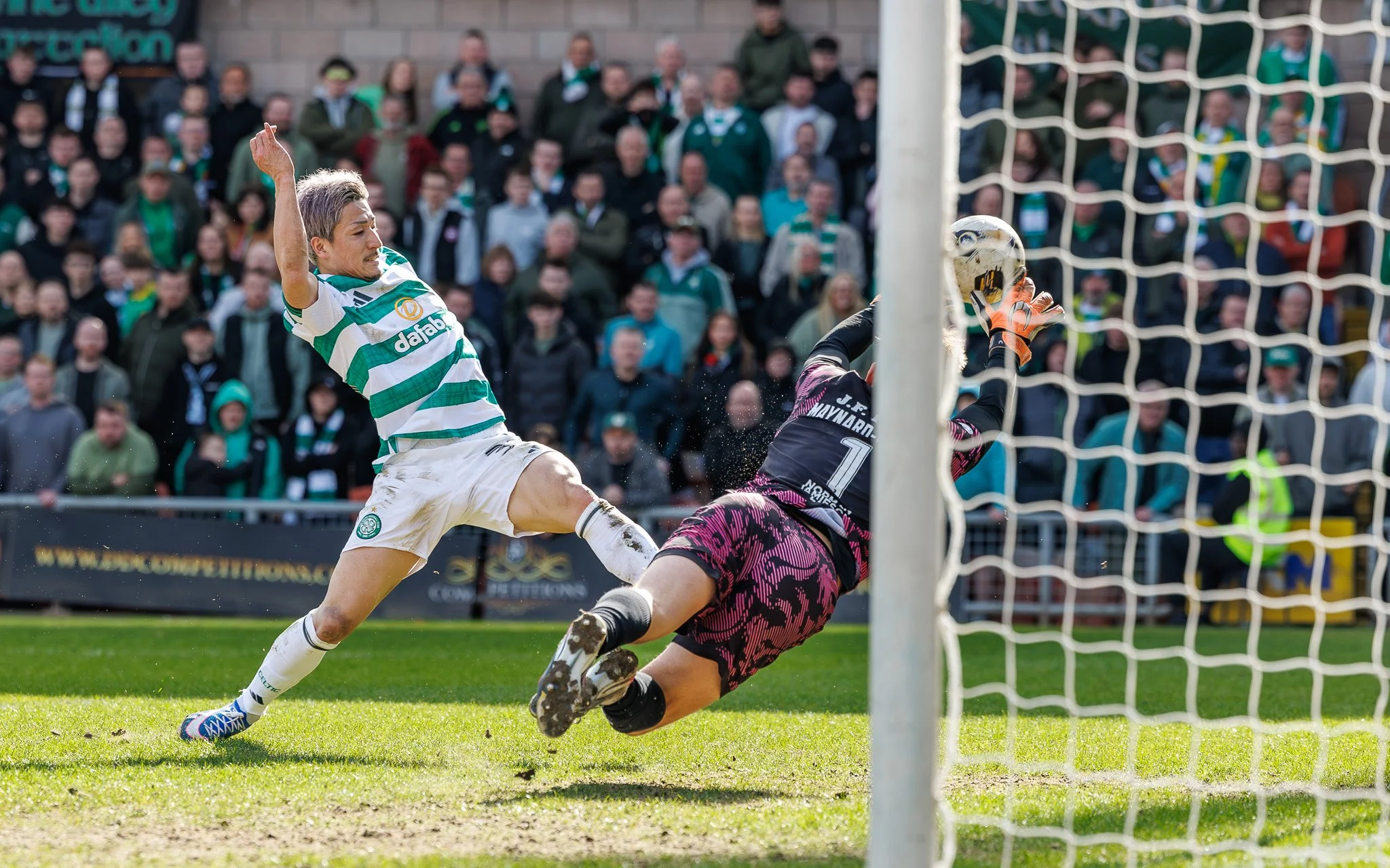 A soccer player in a green and white striped jersey is kicking a ball towards the goal, while a goalkeeper in a pink and black uniform attempts to block the shot, with a crowd of spectators watching in the background.