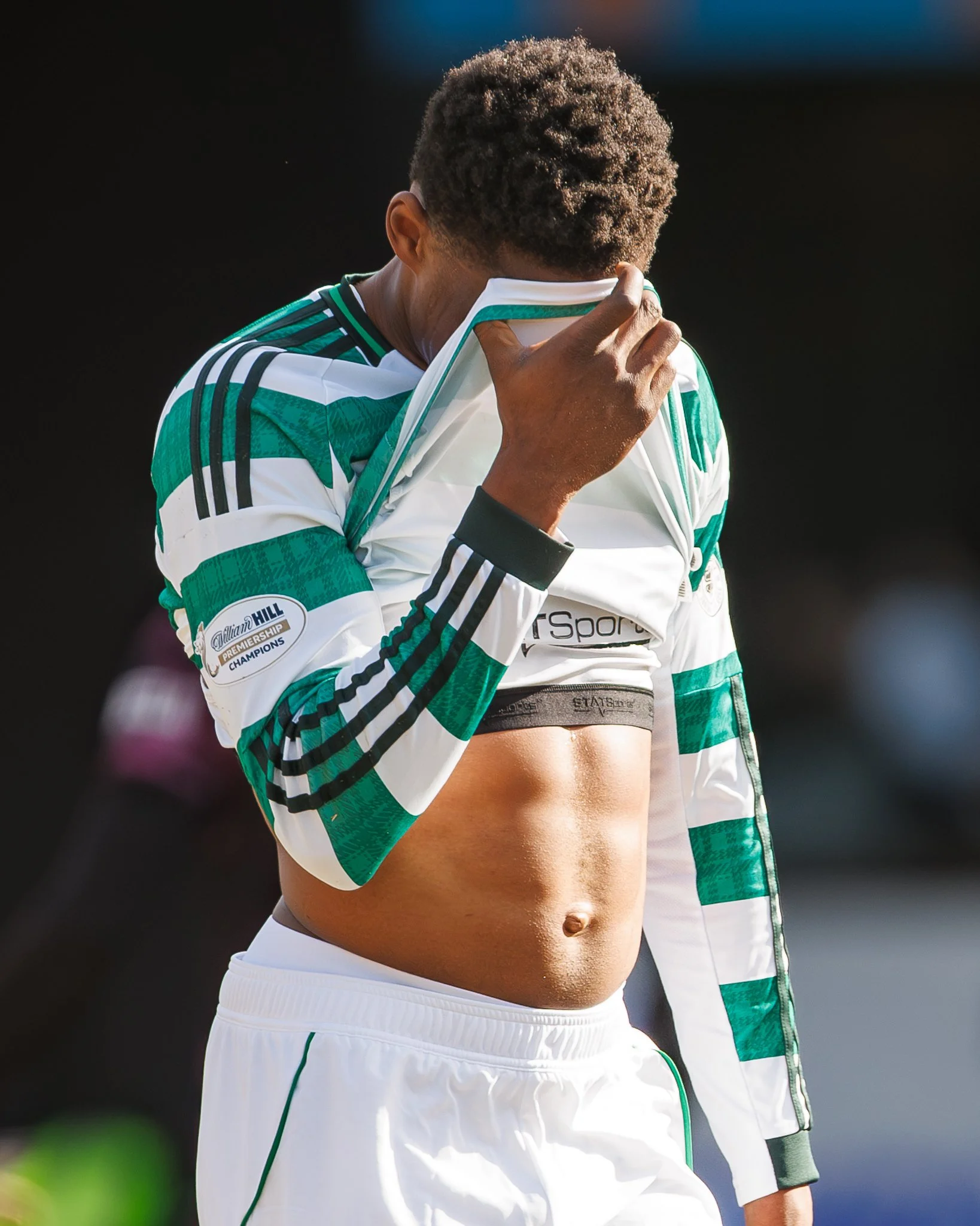 A soccer player in a green and white jersey covering their face with their shirt on a sunny day.