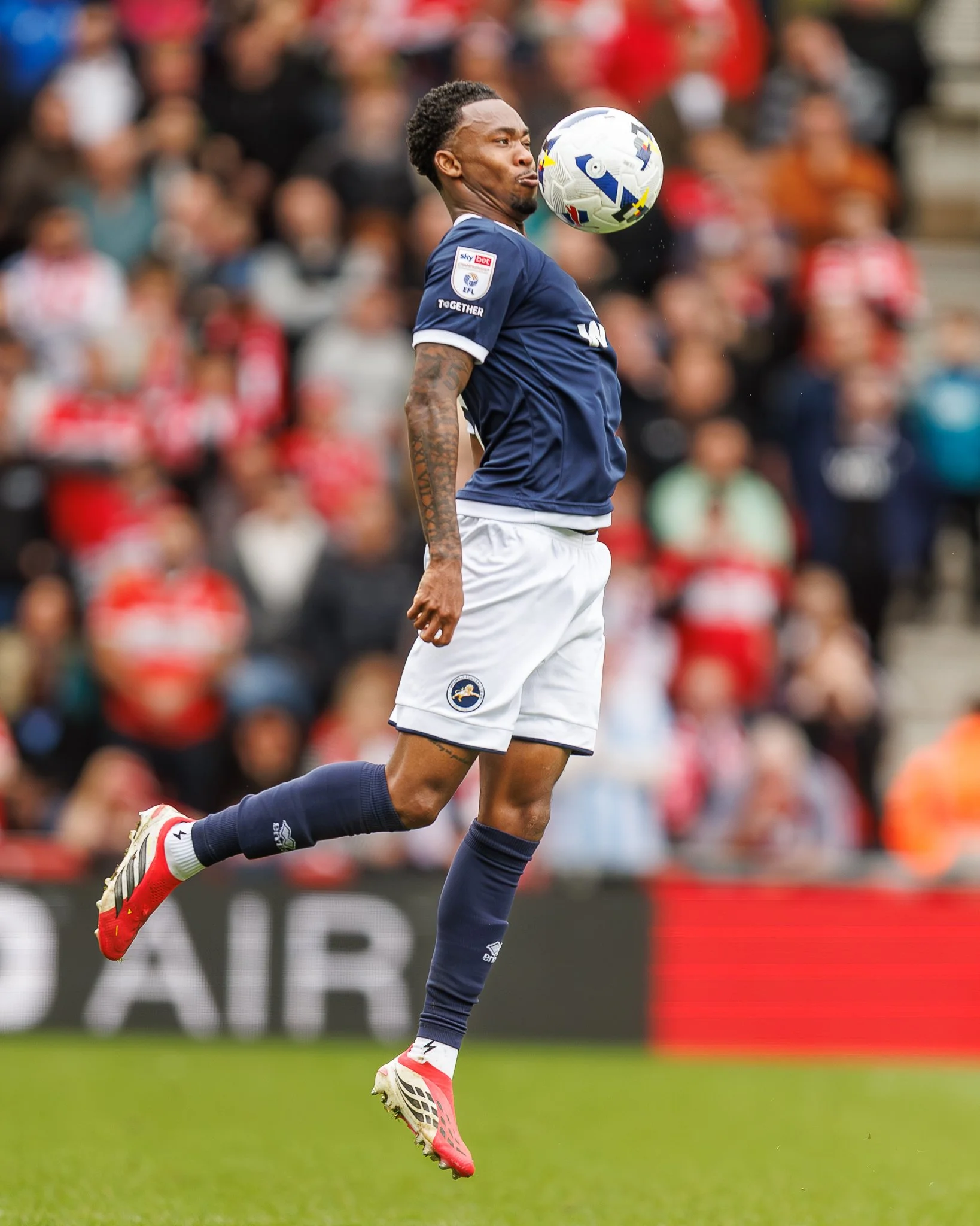 Soccer player in blue jersey jumping to head the ball during a game with spectators in the background.