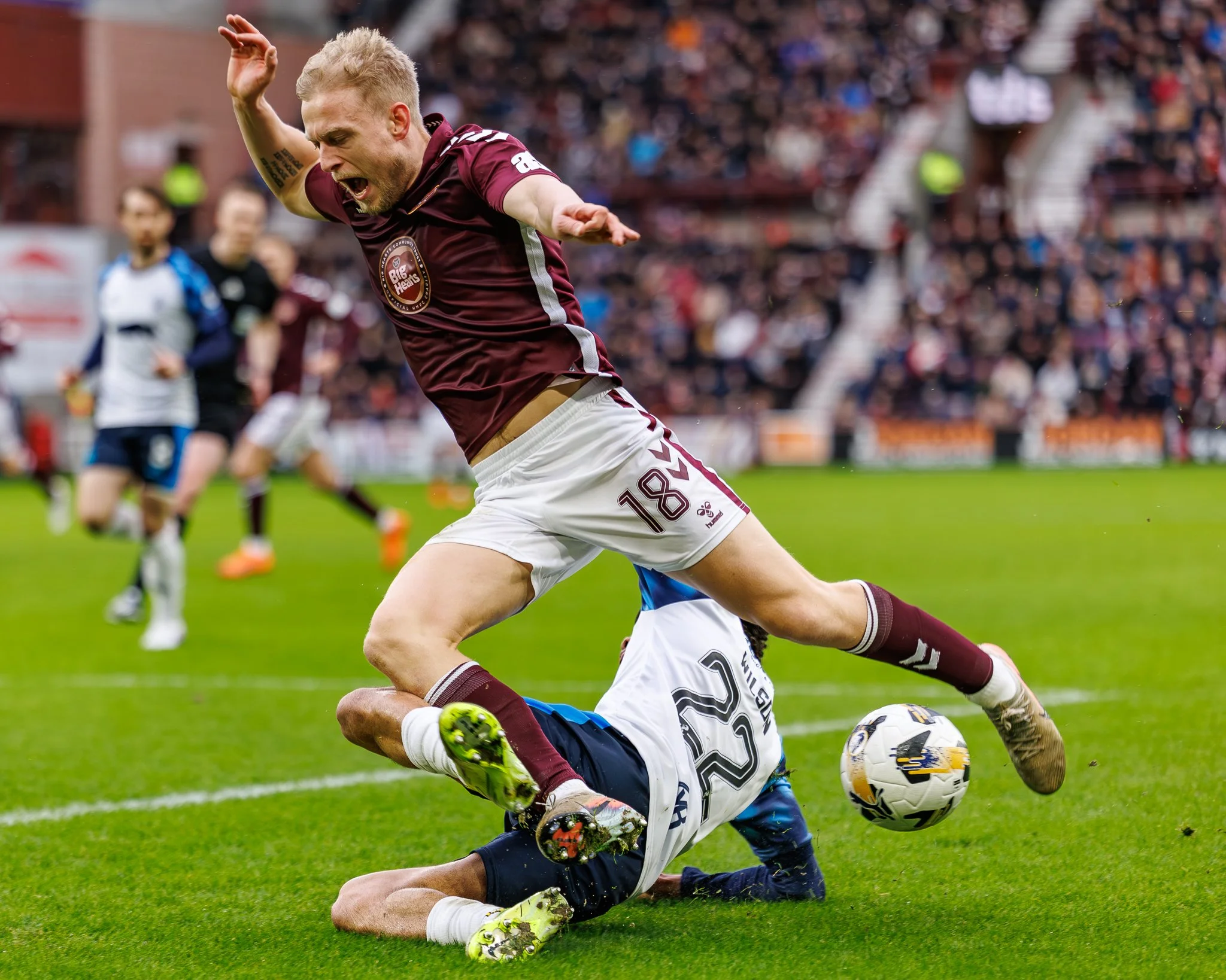 Soccer players in action on the field, with one player in a maroon jersey kicking the ball while another falls.