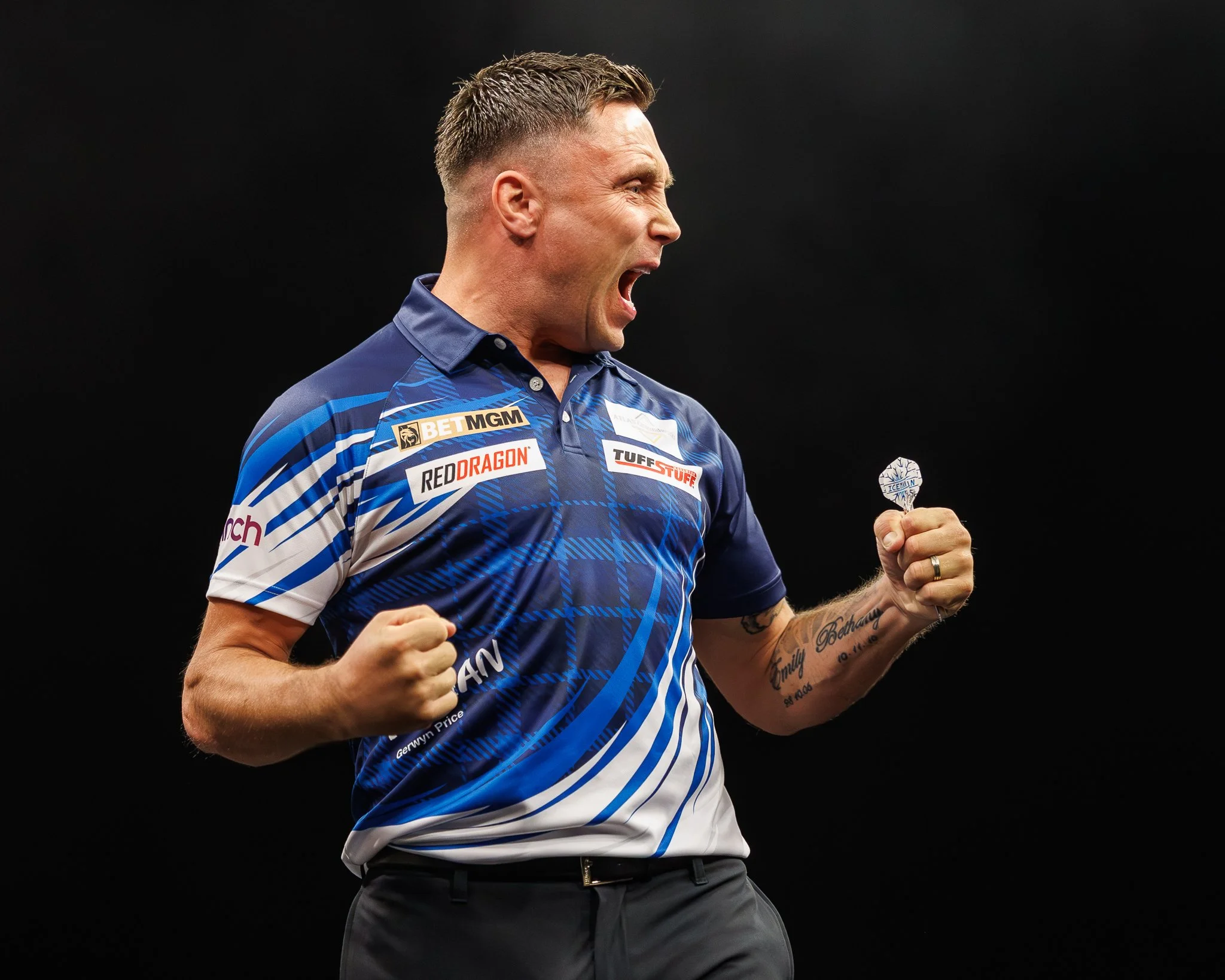 A man wearing a blue sports jersey with sponsor logos, yelling or celebrating, holding a small fan or sign in his left hand, with tattoos on his right arm, against a dark background.