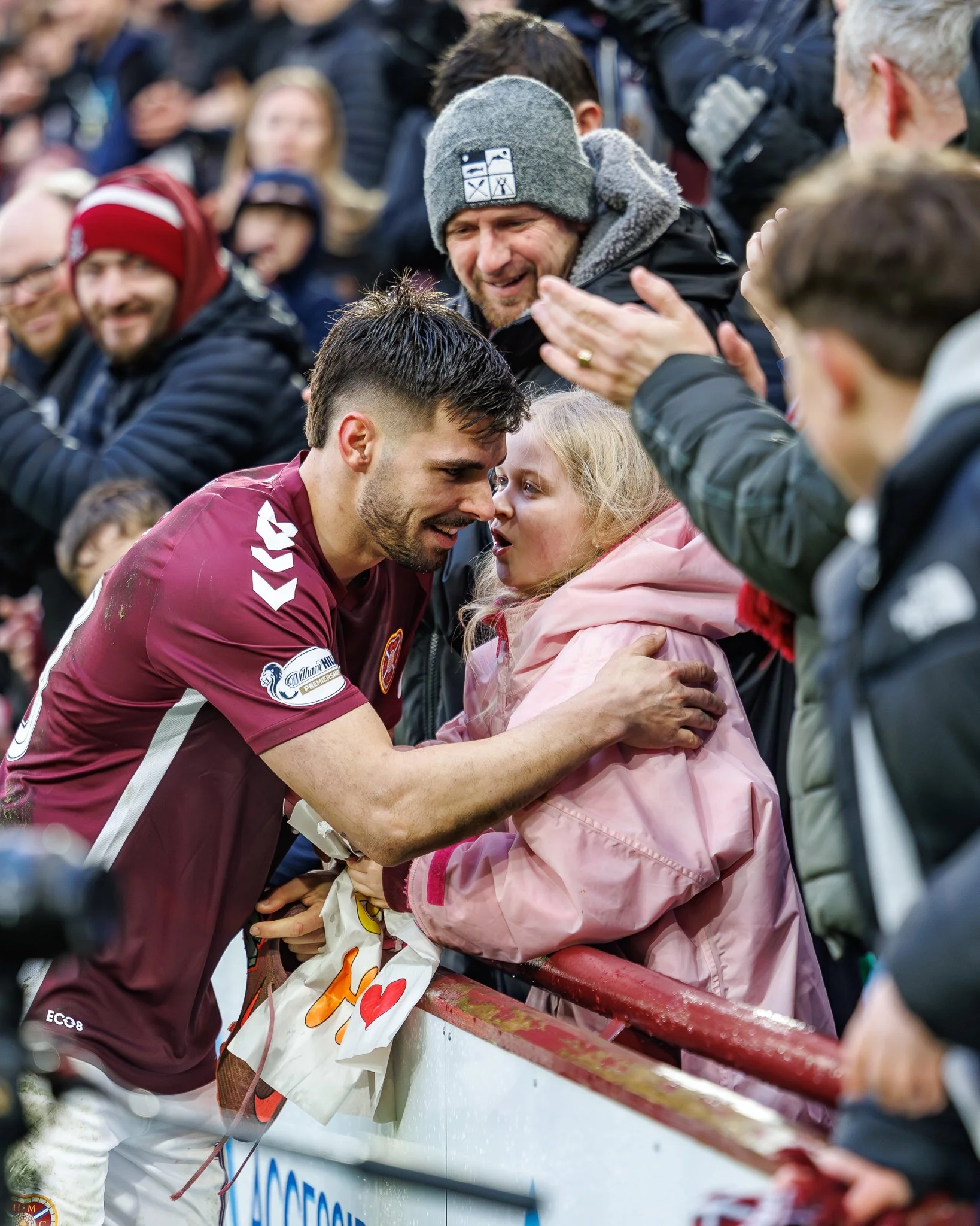 A man and a young girl share a close moment at a sporting event, with the man holding a painted sign reading 'I ♥' and a partially visible bottle of soda. The background shows a crowd of spectators.