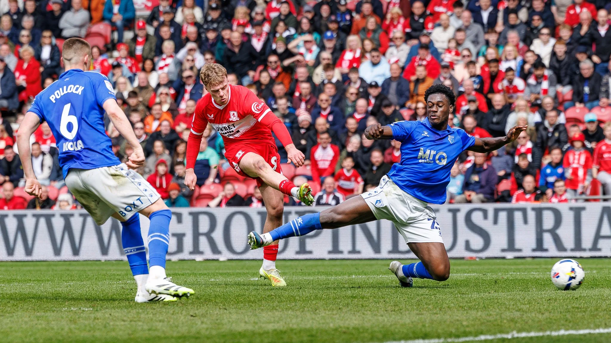 Three soccer players in action during a match, with one player kicking the ball while two others move around him. Spectators watch from the stands in the background.
