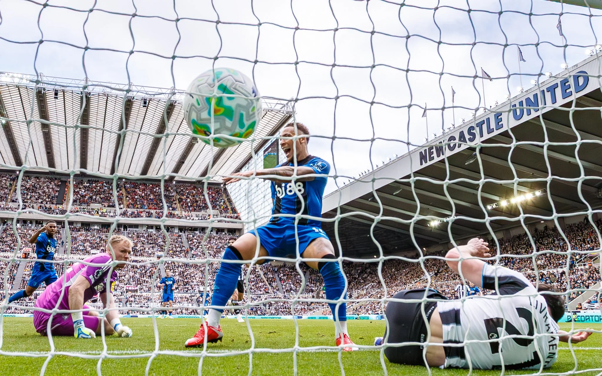 Soccer players and a goalkeeper on the field during a match, viewed through the goal net, with a large crowd in the stadium and a sign that reads 'NEWCASTLE UNITED' in the background.