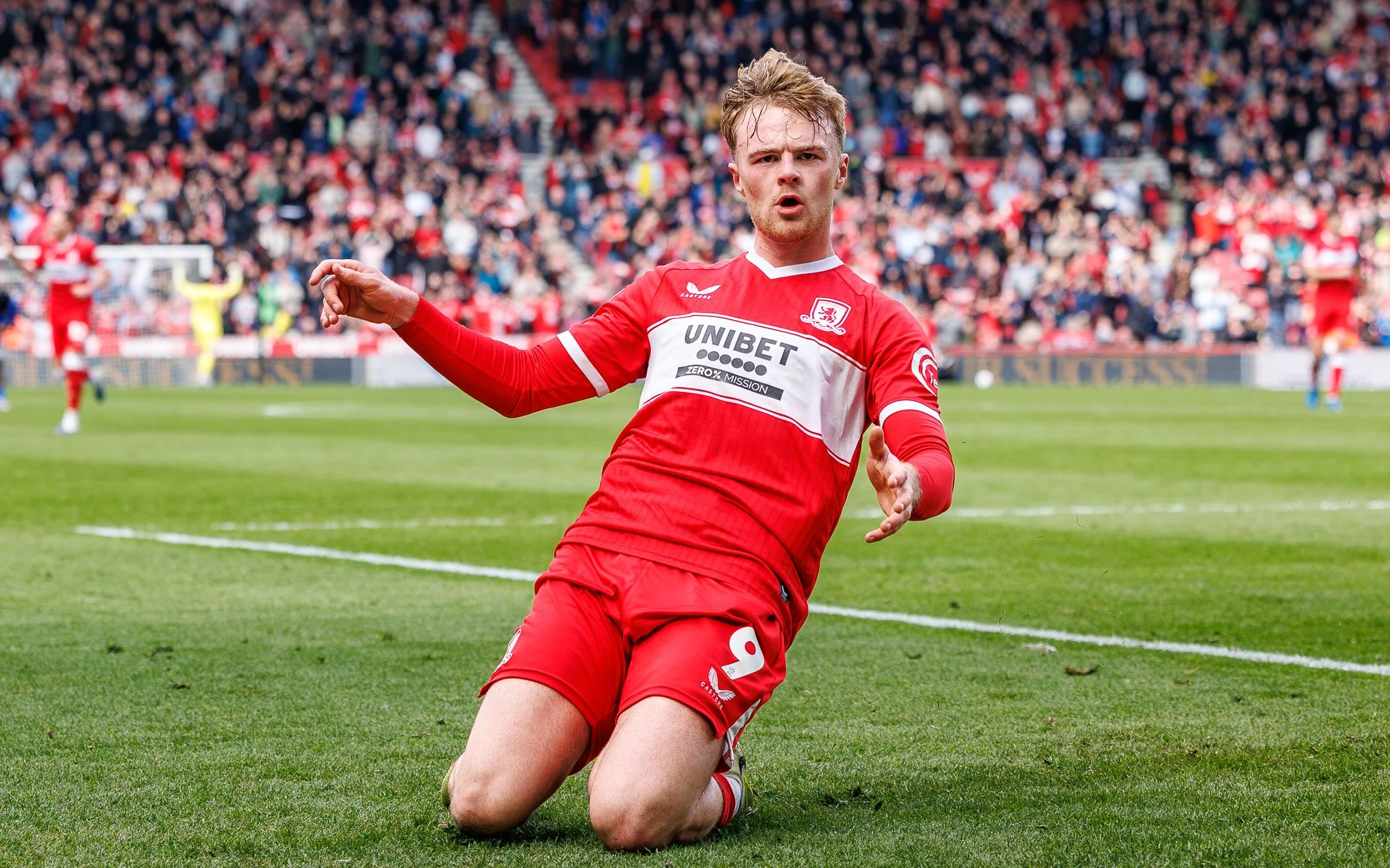 A soccer player in a red uniform is kneeling on the field with a surprised or expressive face. He is in front of a large crowd in the stands.