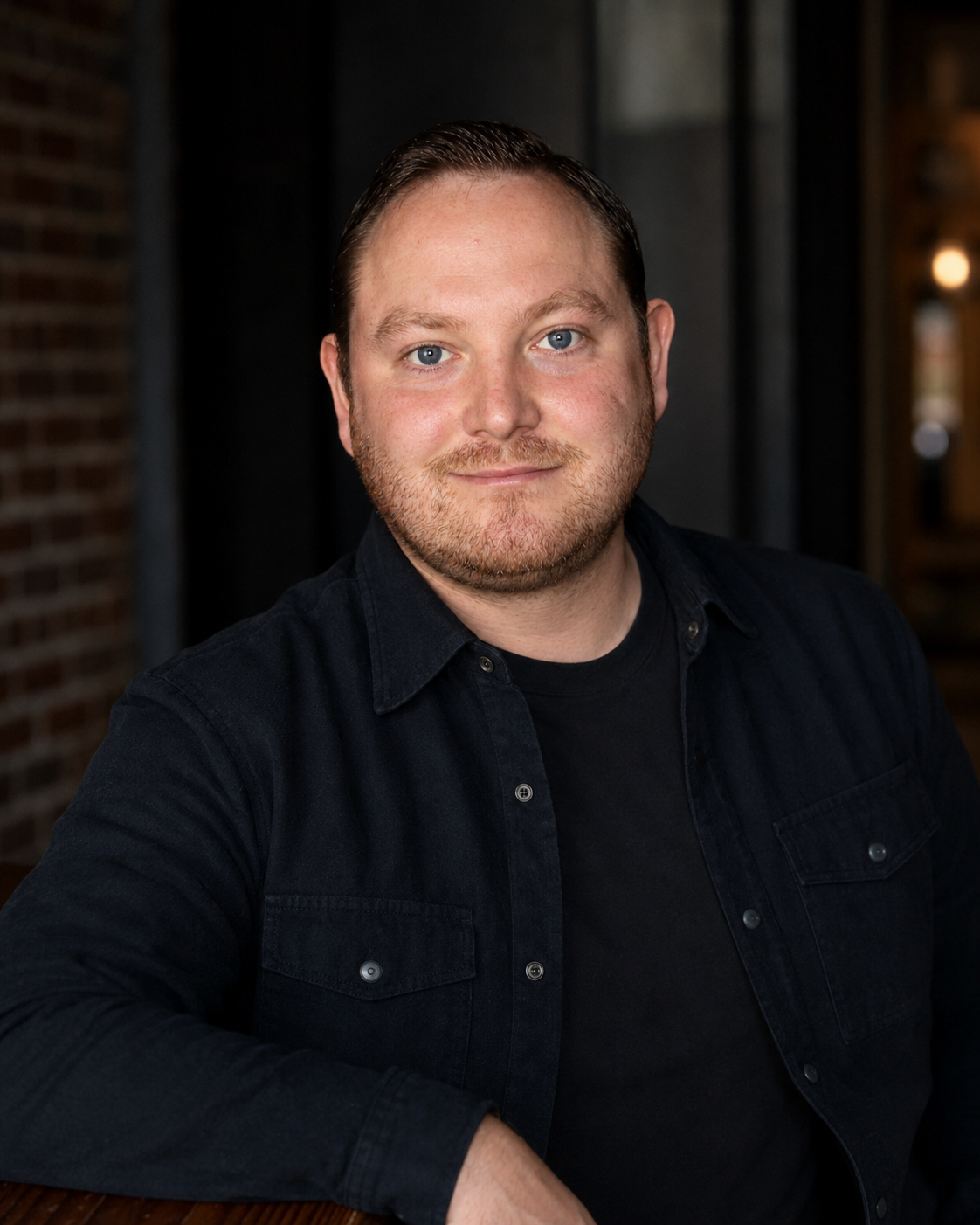 A man with light skin, blue eyes, and short brown hair, wearing a black shirt, sitting indoors with a brick wall and dark curtains in the background.