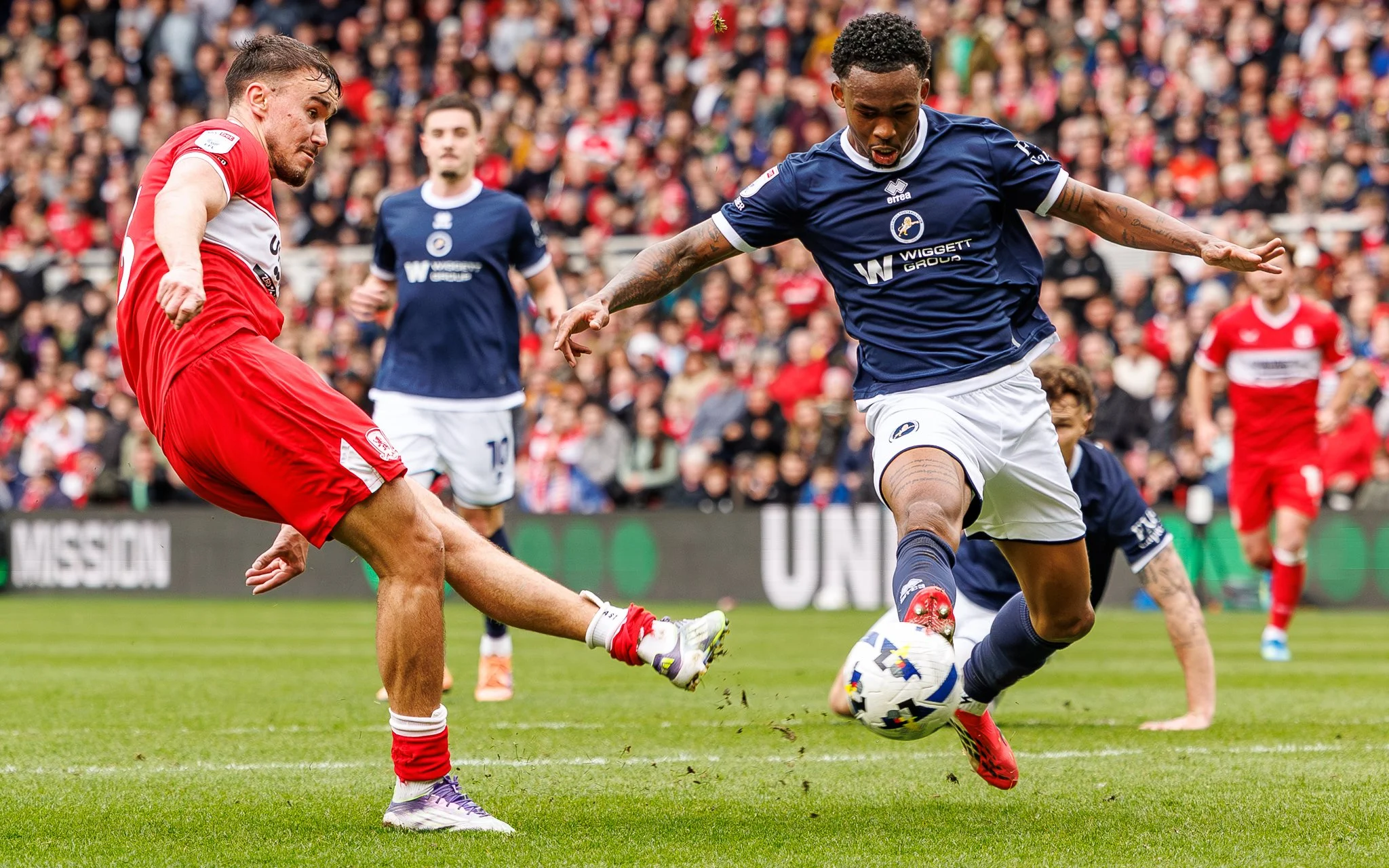 Two soccer players are competing for the ball during a match, with a crowd in the background.