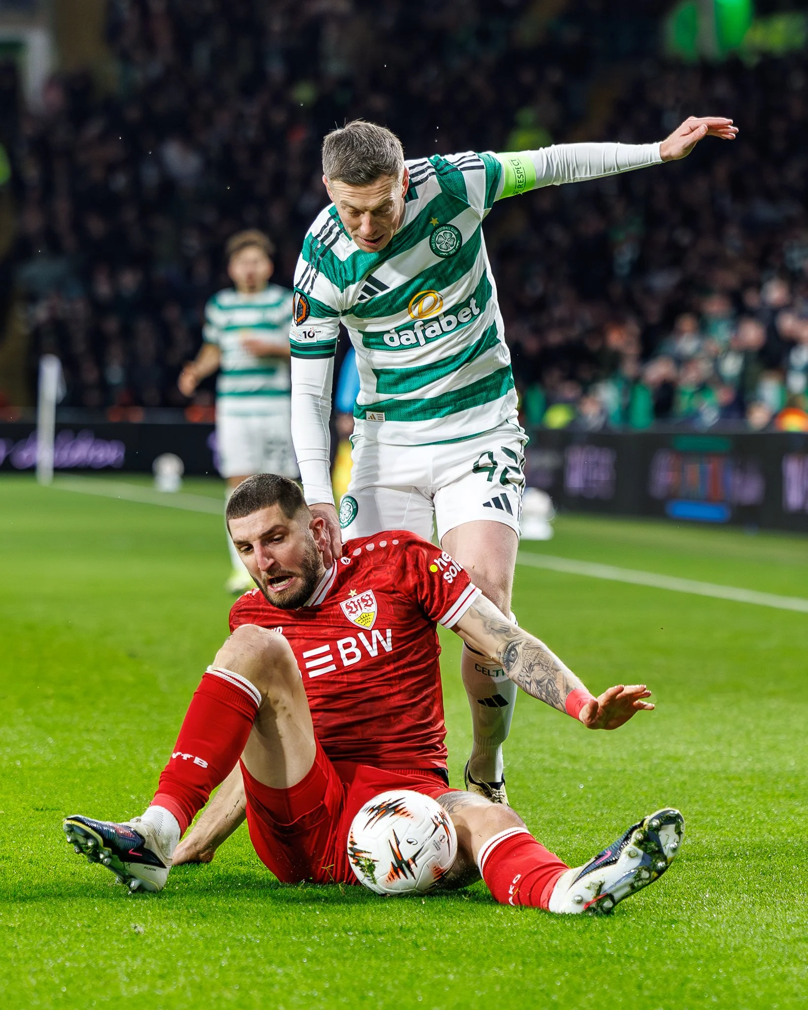 Two soccer players are competing for the ball on the field during a match. One player in a red uniform is sitting on the ground, holding the ball, while the other player in a green and white striped uniform is leaning over him. The background shows a