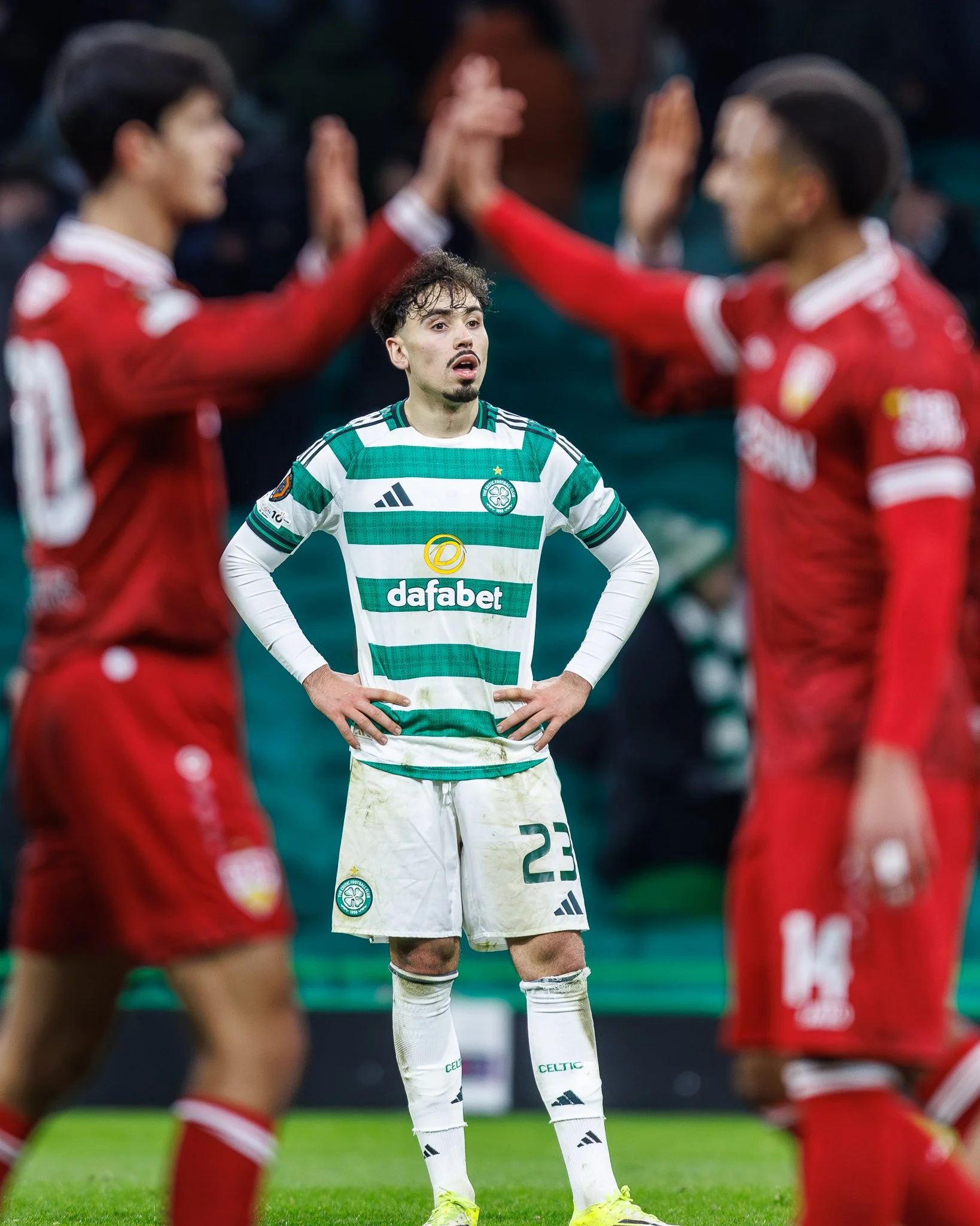 A soccer player in a green and white striped jersey stands with hands on hips, while teammates in red uniforms are congratulating each other in the background.