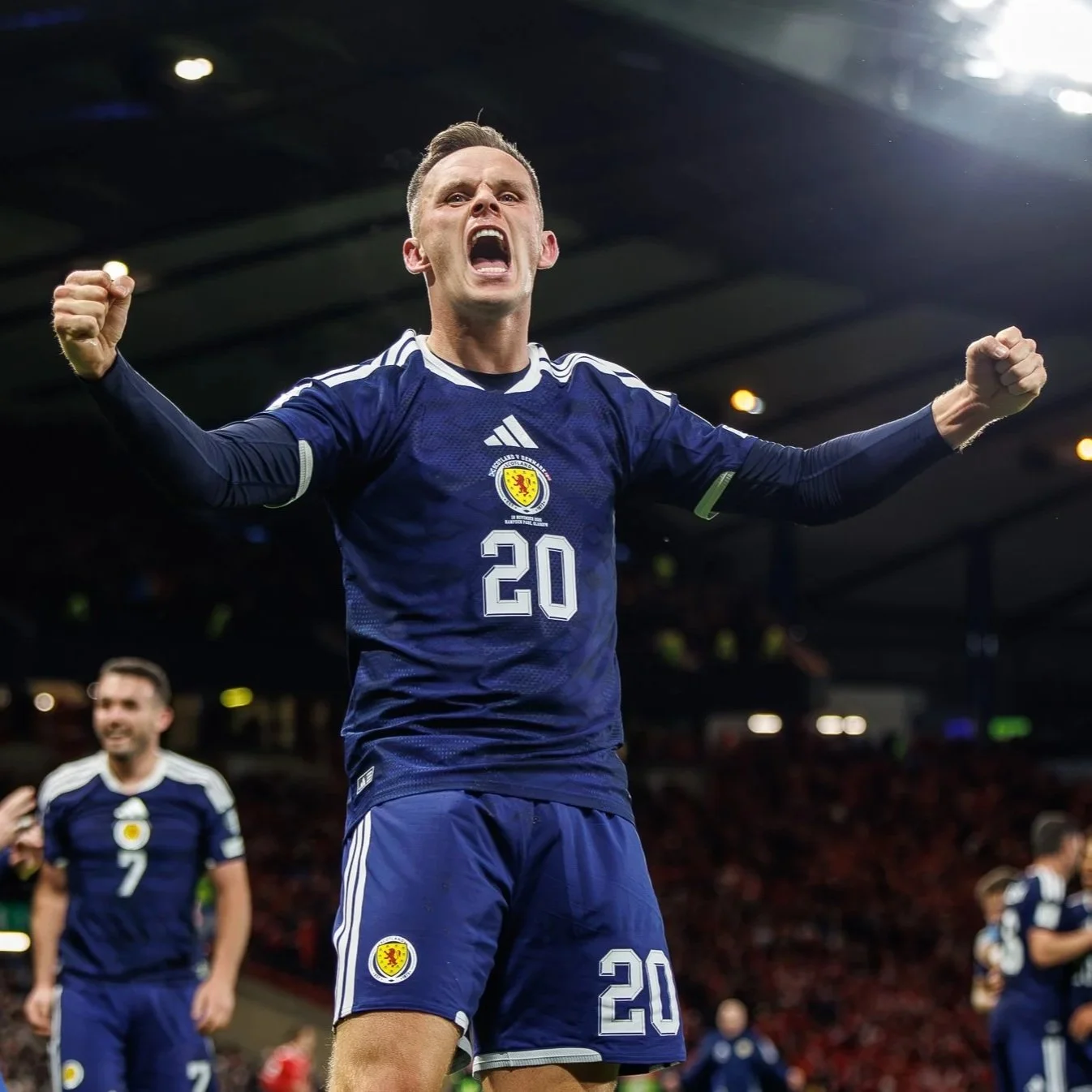 A soccer player celebrating on the field, wearing a blue jersey with the number 20 and a crest. He is raising his fists and appears to be shouting in excitement. Other players and a stadium are visible in the background.