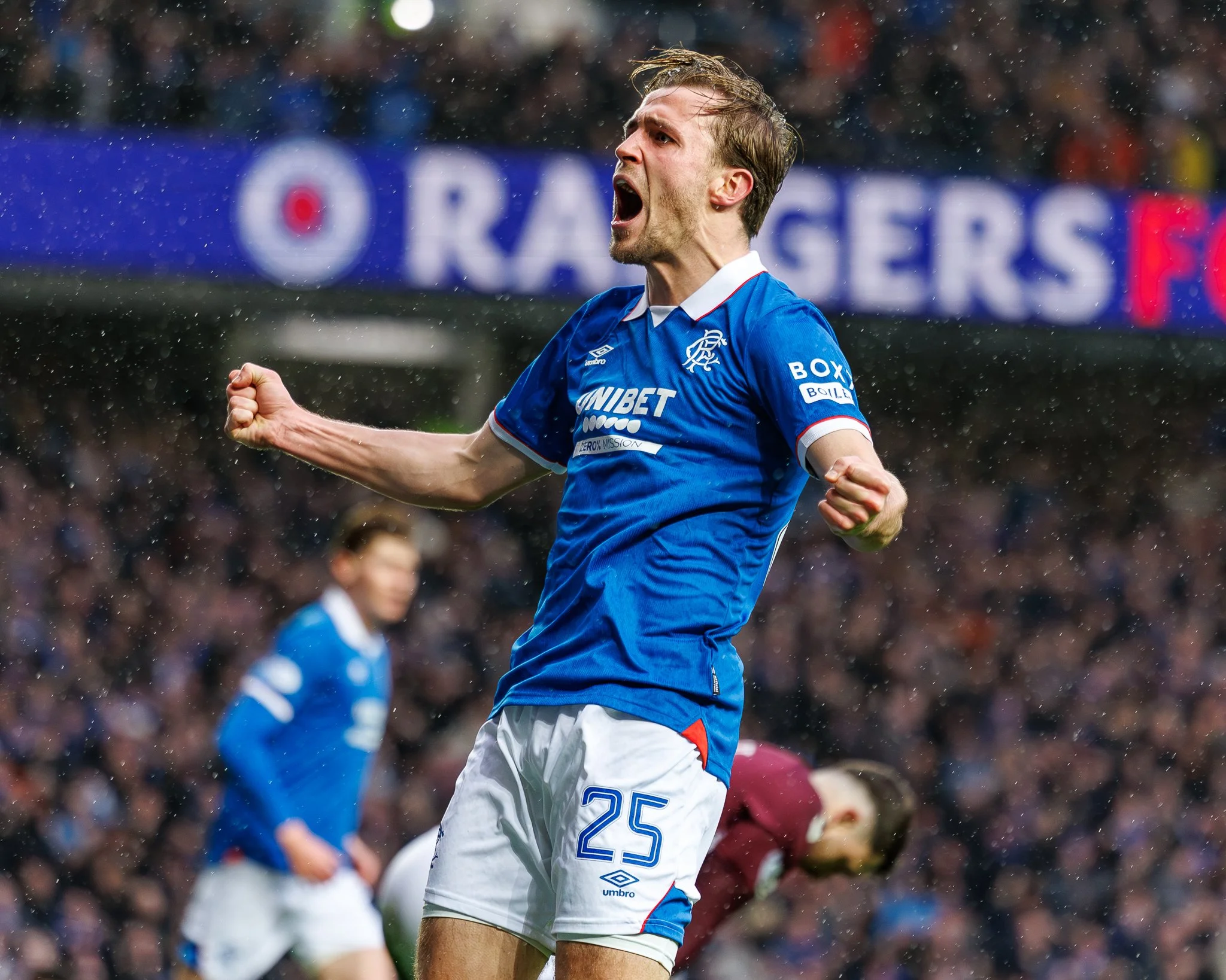 A male soccer player with short brown hair wearing a blue jersey celebrating on a soccer field in the rain, with his fists clenched and mouth open.