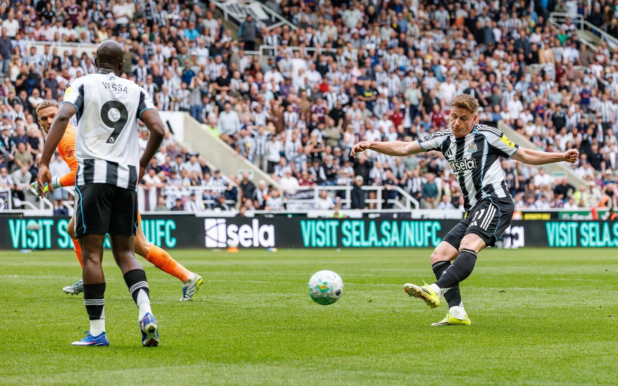 Soccer players in black and white jerseys compete for the ball on a grass field during a match, with a crowded stadium in the background.