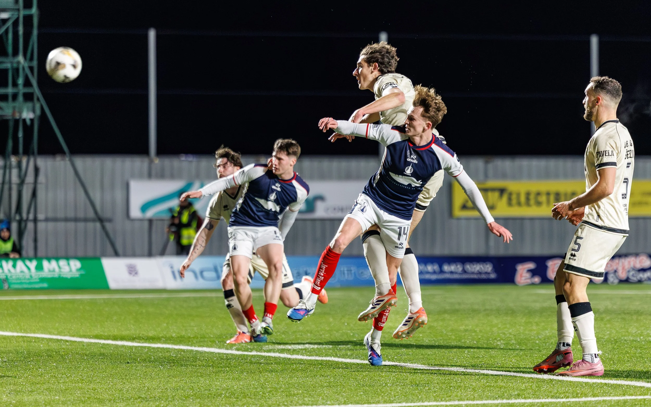 Soccer players competing for the ball during a match at night, with some players jumping and others on the ground, on a lit field with advertising banners in the background.