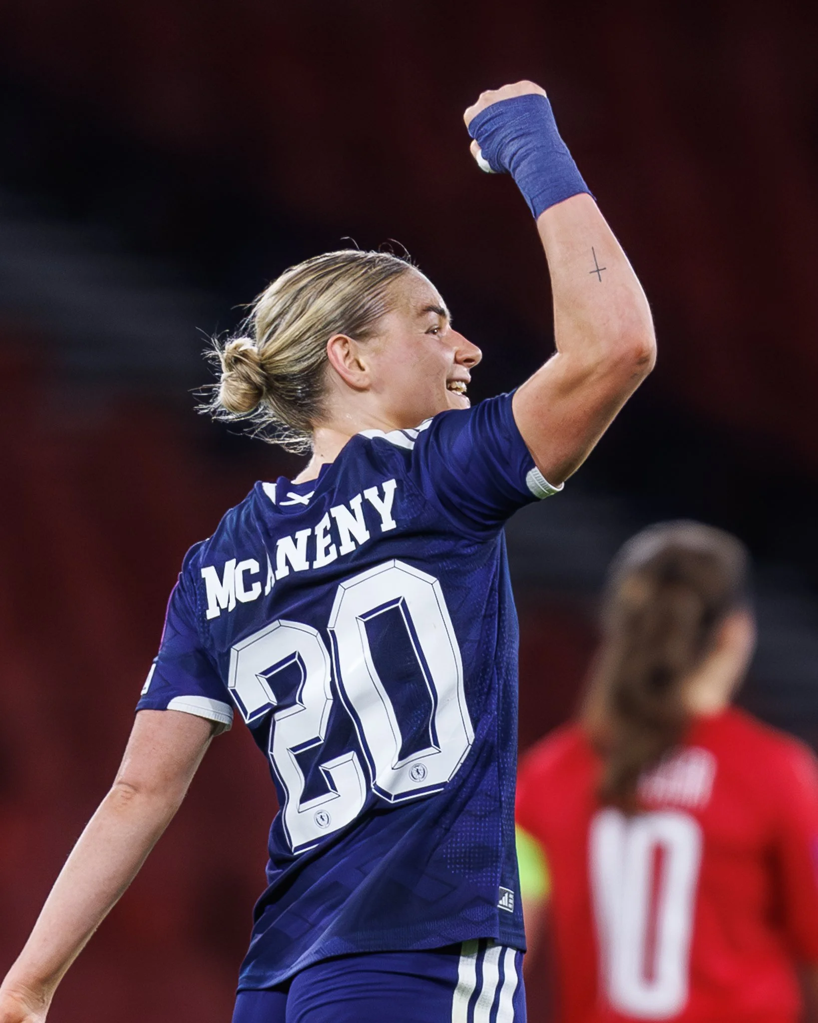A female soccer player with blonde hair tied in a bun, wearing a blue jersey with the name "McEnany" and the number 20, raising her arm in celebration on the field during a match.