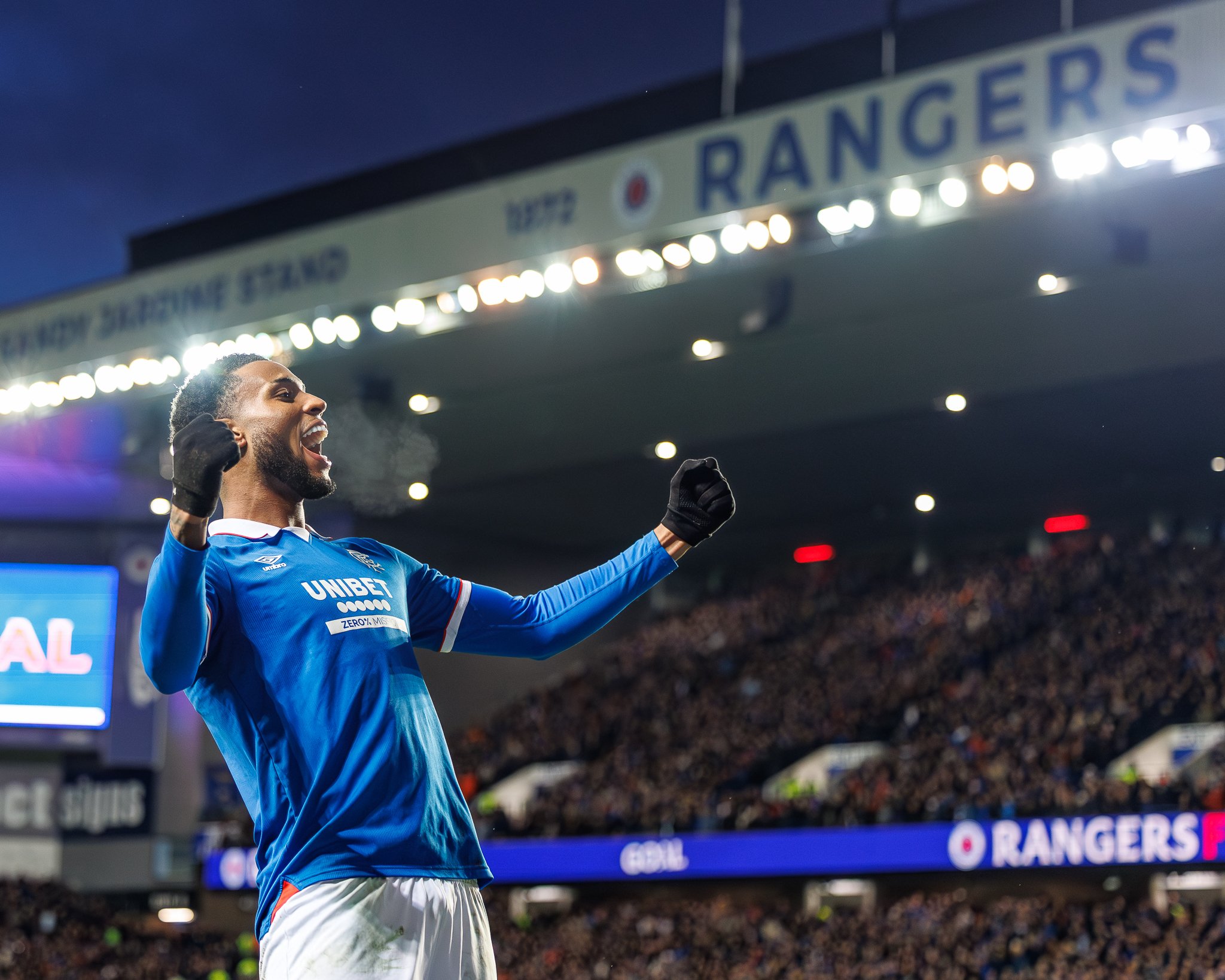 A soccer player in a blue jersey celebrating on the field at night, with a stadium filled with spectators in the background and a sign that reads 'RANGERS' overhead.