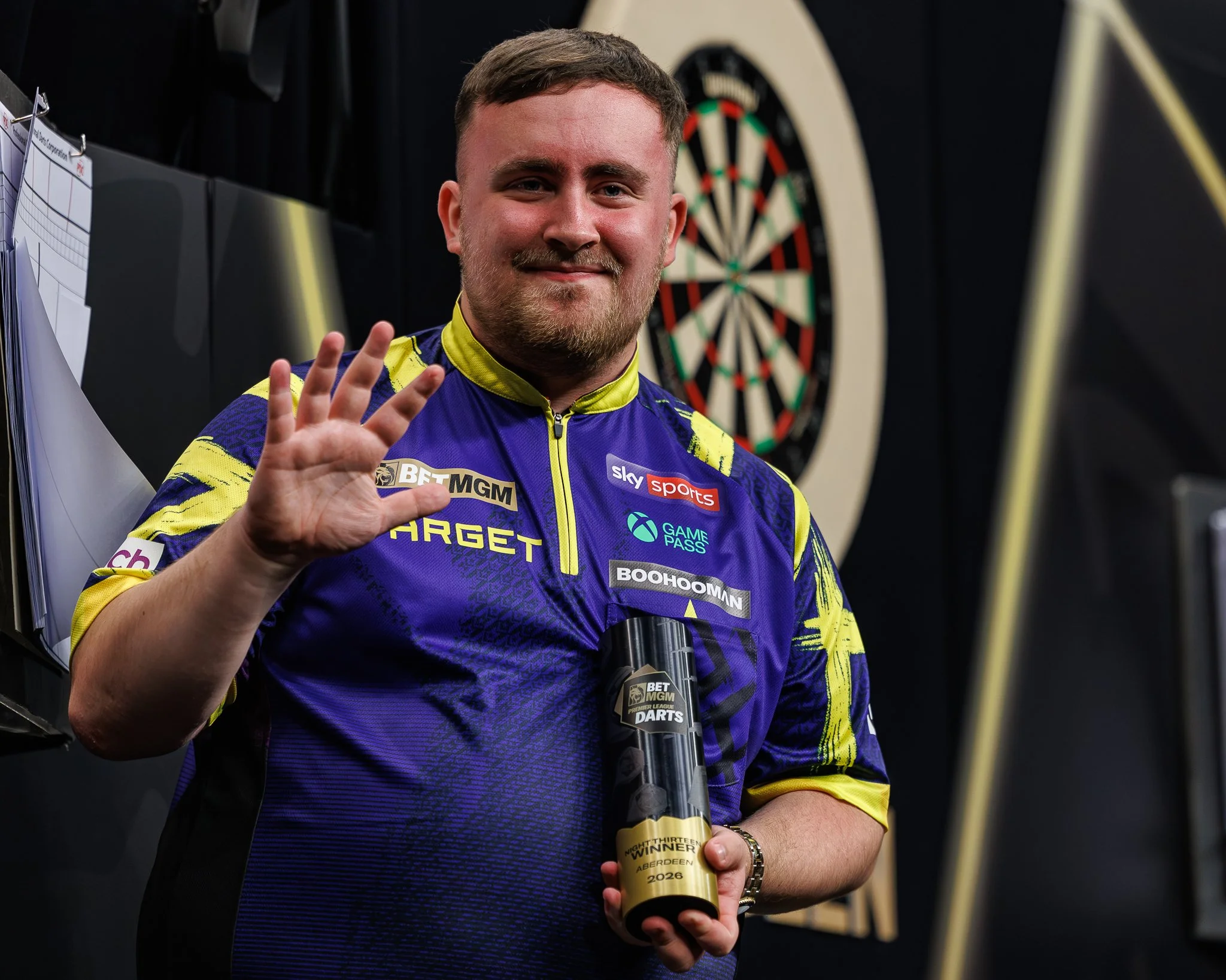 A man in a yellow and purple esports jersey is smiling and holding a trophy in front of a dartboard. The trophy has a label that says 'NIGHTFRIGHT WINNER ABERDEEN 2020 2026.'