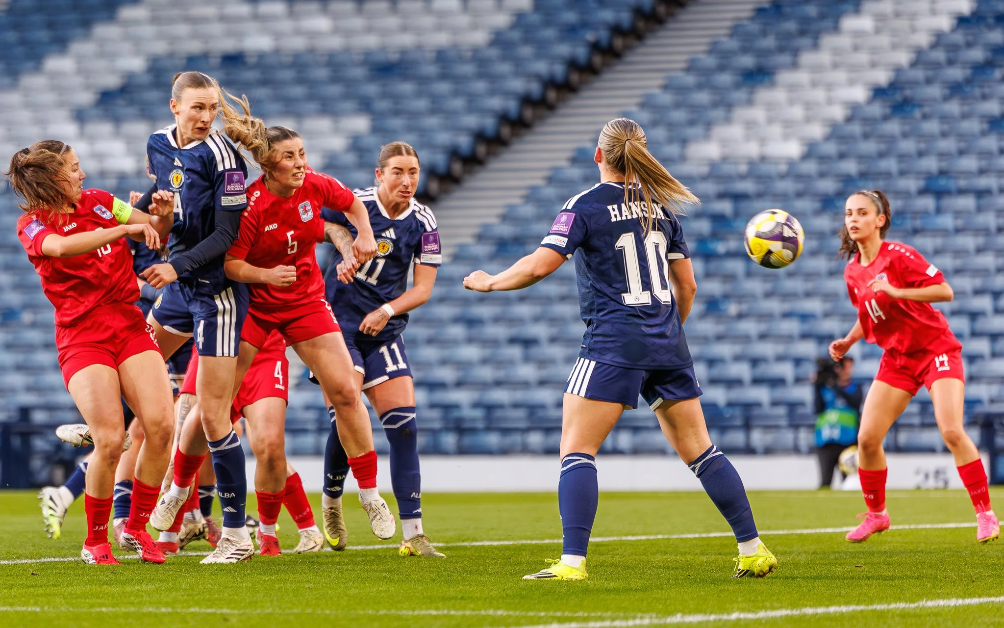 Women soccer players in blue and red uniforms competing for the ball on a soccer field in a stadium.