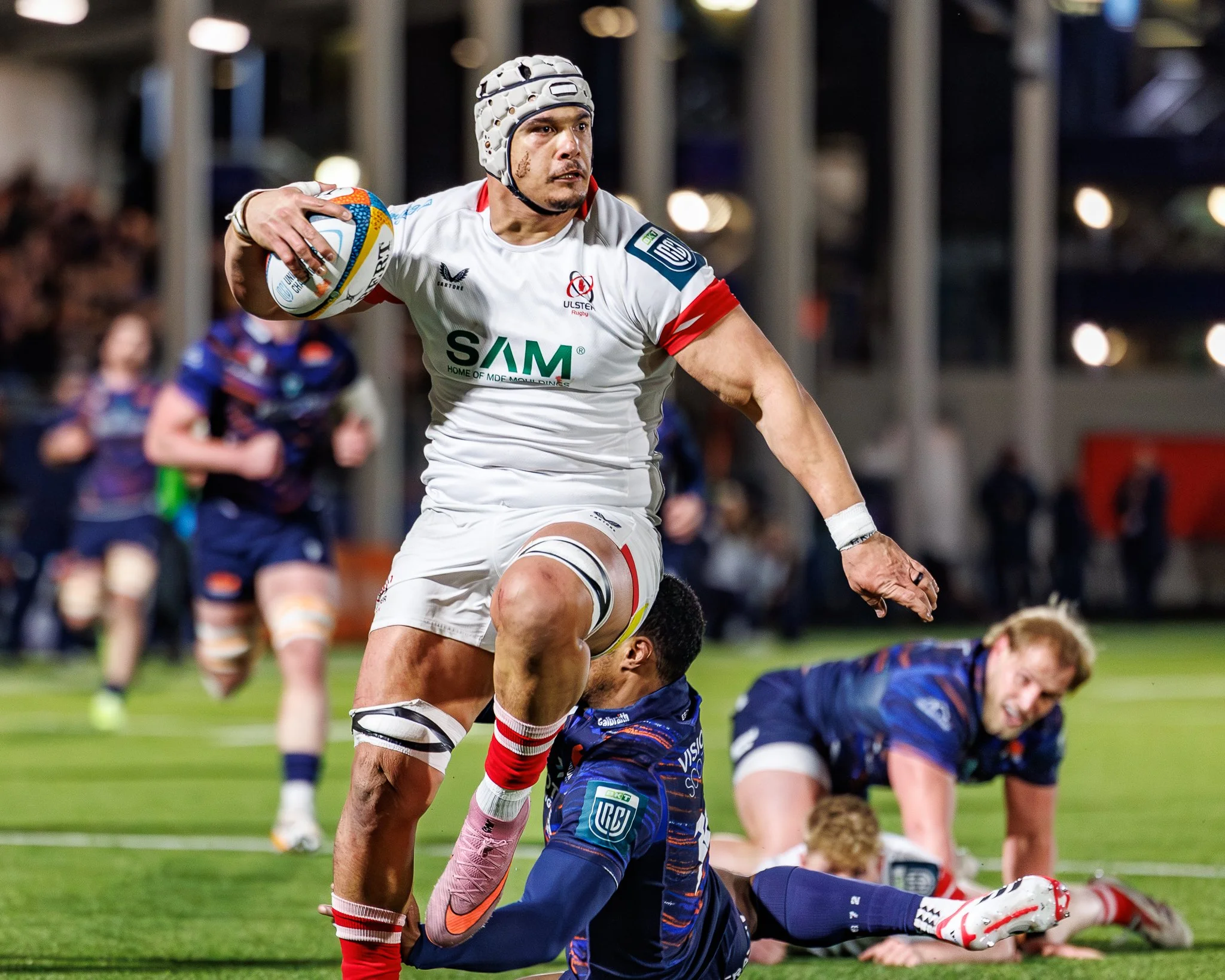 A rugby player in a white and red uniform holding a rugby ball while running on the field, with other players in blue uniforms on the ground and blurred spectators in the background.