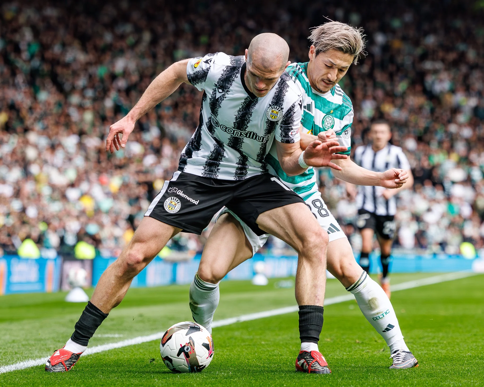 Two soccer players from opposing teams competing for the ball on the field, surrounded by a crowd of spectators in the background.