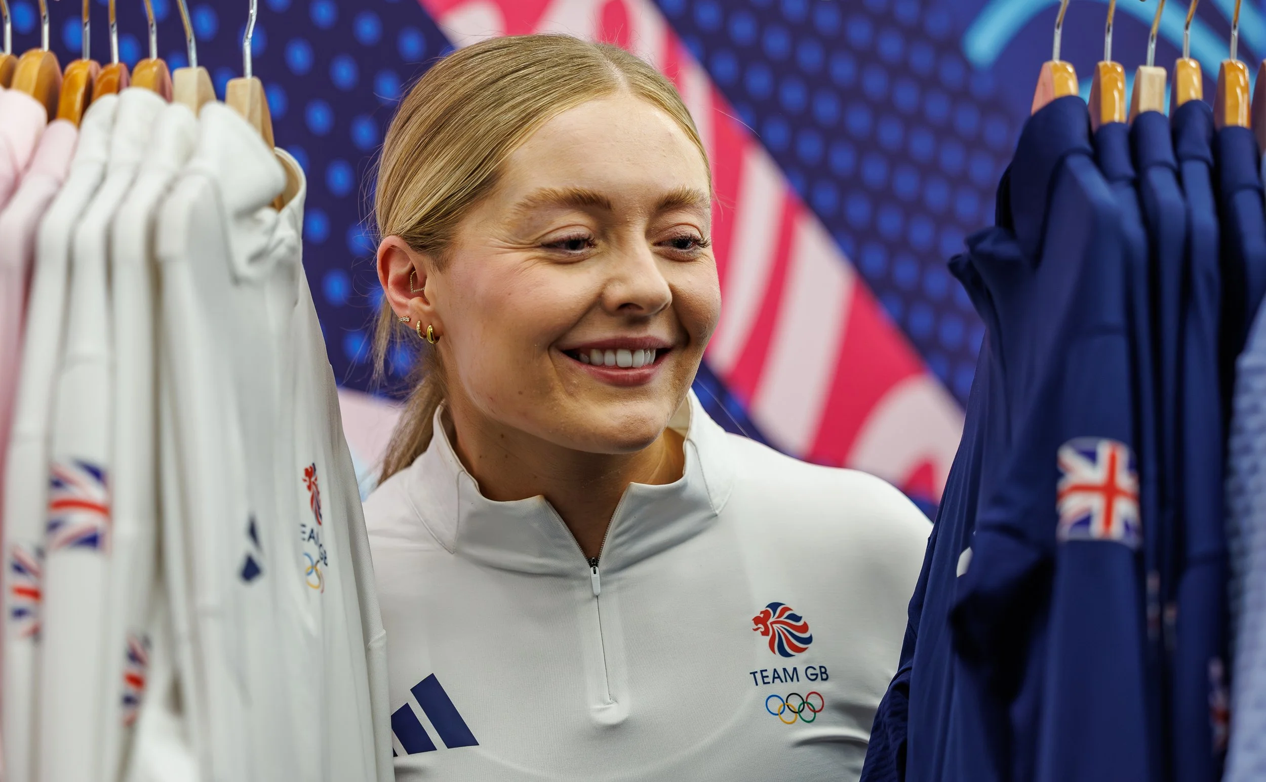 Close-up of a smiling female athlete with blonde hair, wearing a white jacket with the British flag, Olympic rings, and 'TEAM GB' logo, looking at sports uniforms on racks in the background.