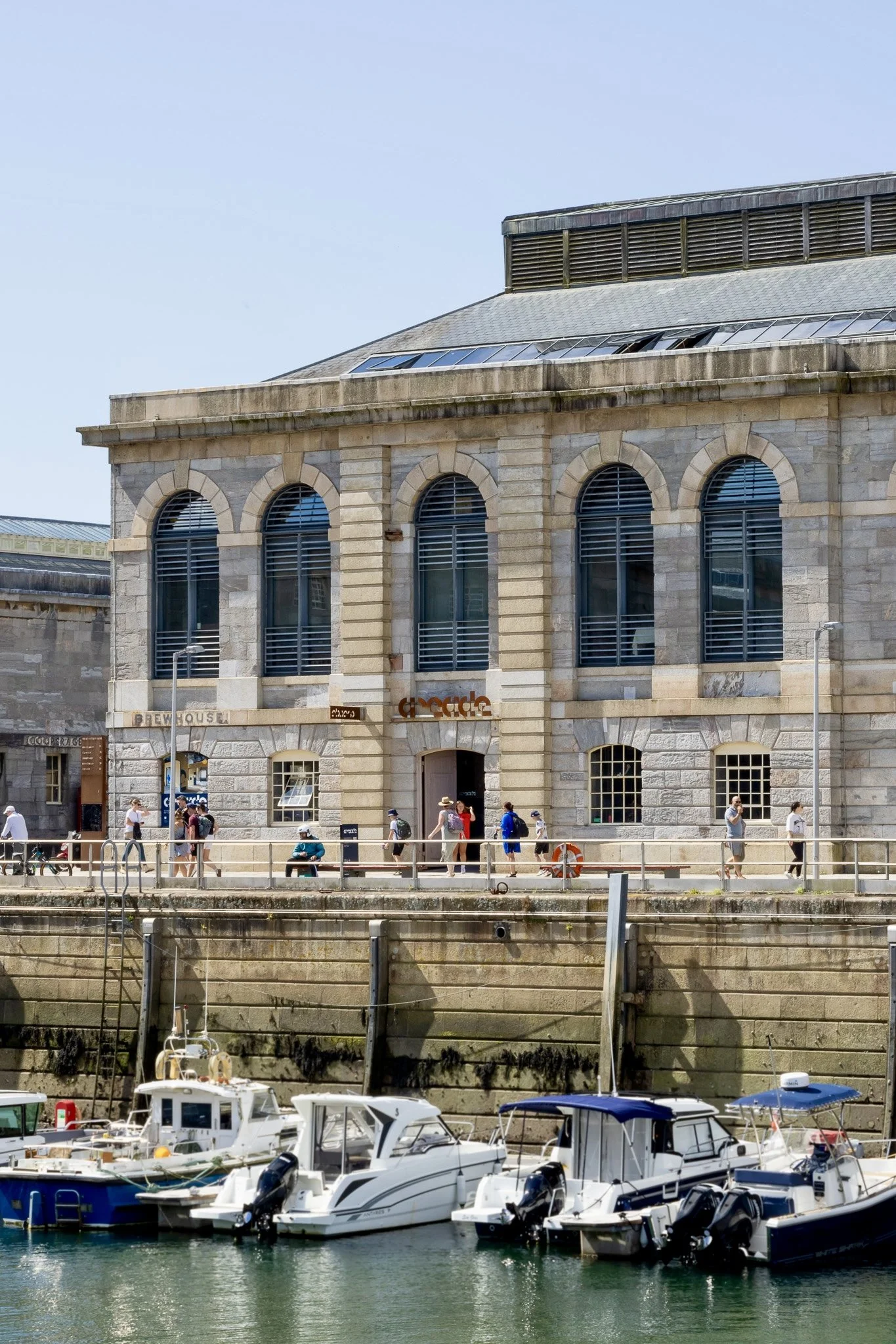 Boats docked along a marina outside of Arcade building, with a historic brick building and people walking along the waterfront.