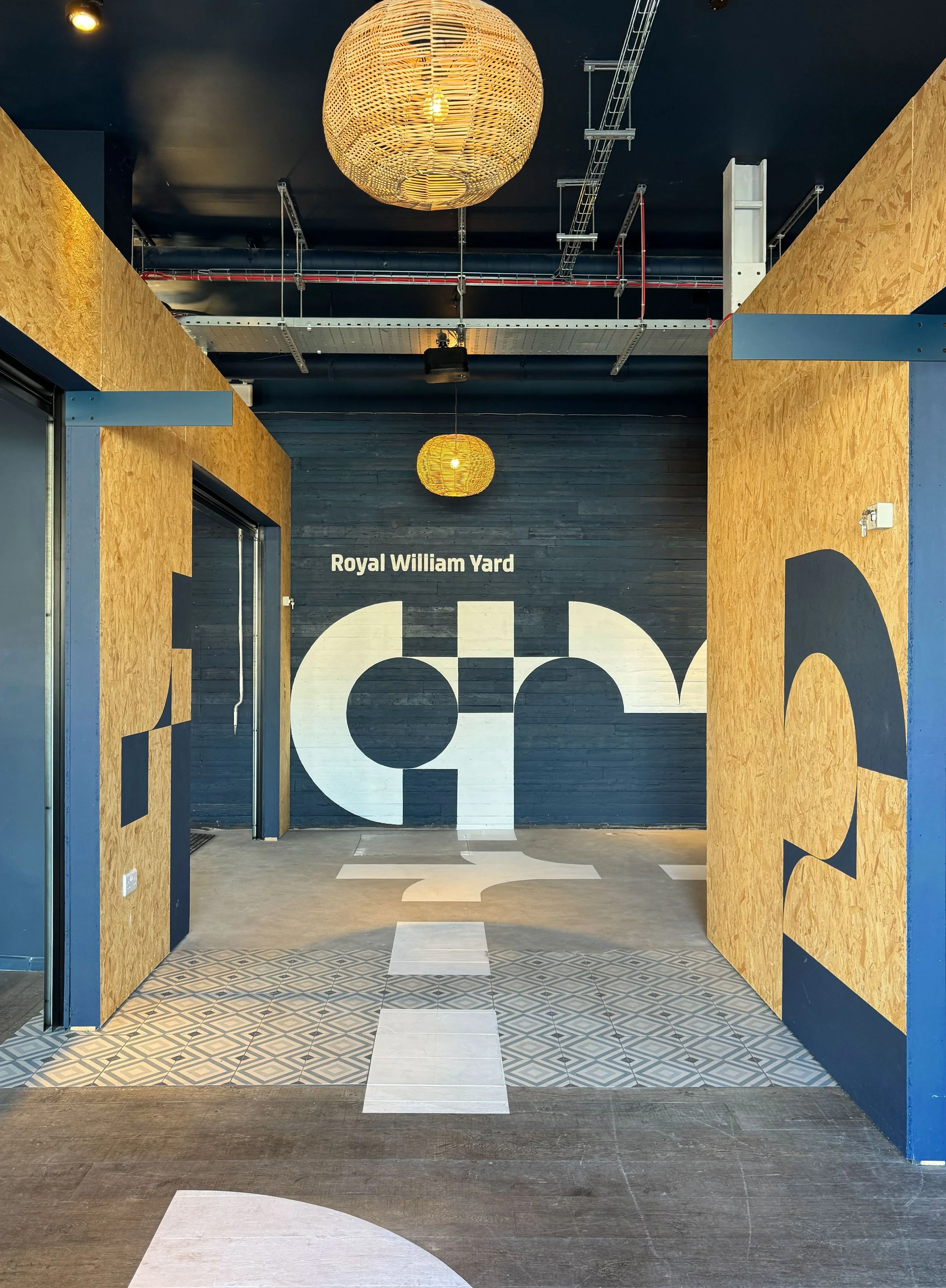 Interior of Arcade, a modern space with wooden partitions, a dark wall with the words 'Royal William Yard,' decorative pendant lights, and patterned floor tiles.