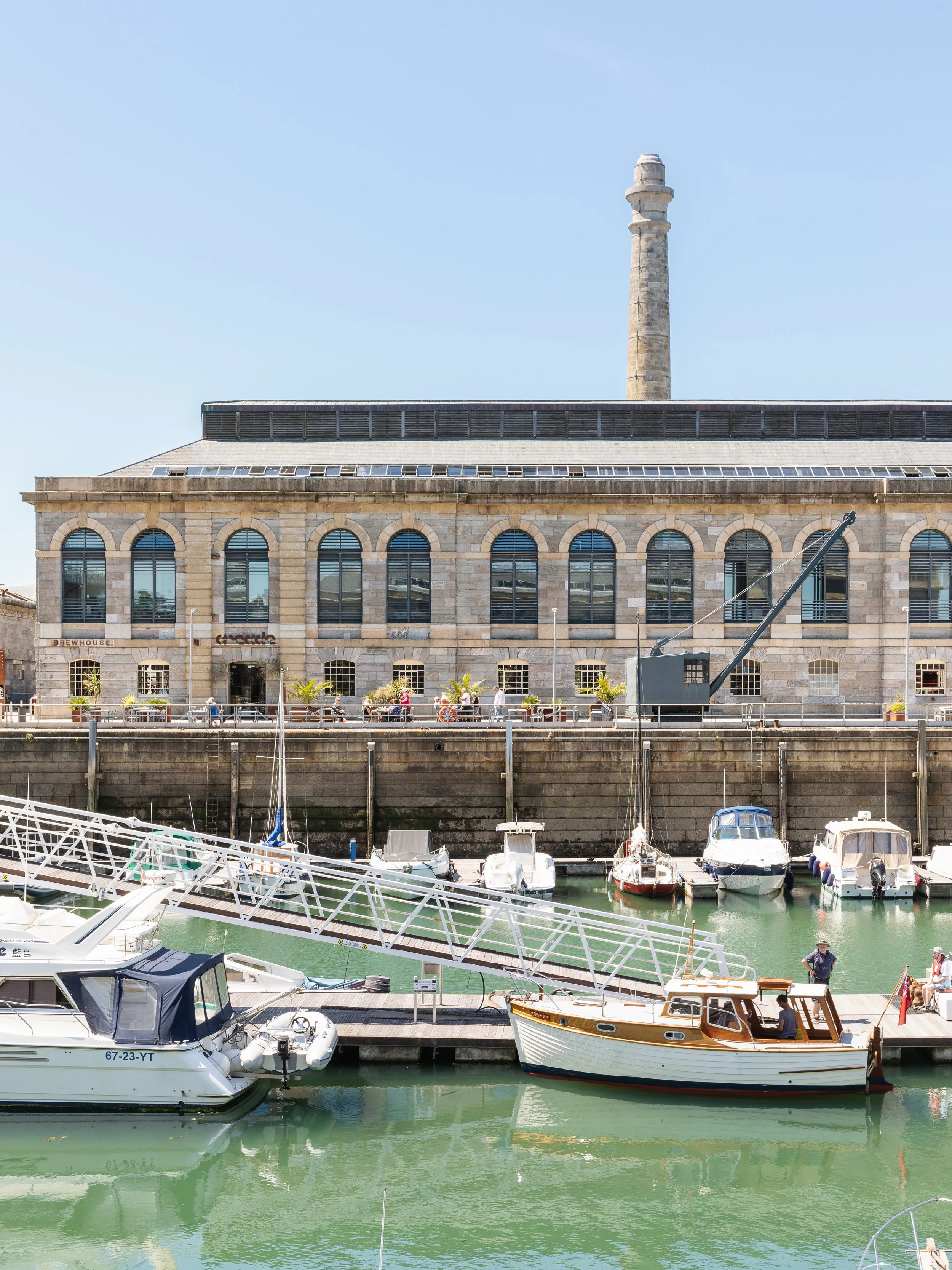 A harbor scene with boats docked in calm water, a historic stone building with arched windows and a chimney in the background, and people walking along the waterfront.