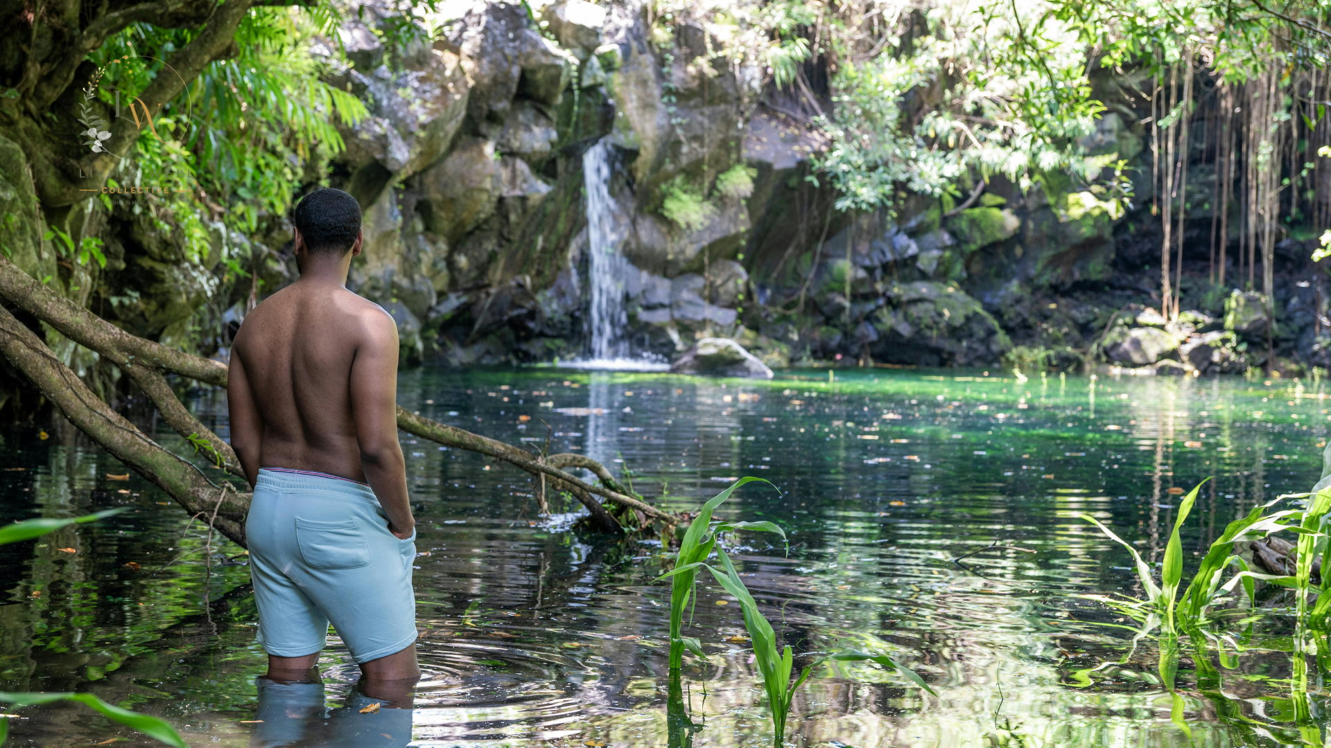 A shirtless man wearing light blue shorts stands knee-deep in a river, facing a small waterfall surrounded by lush green trees and rocks.