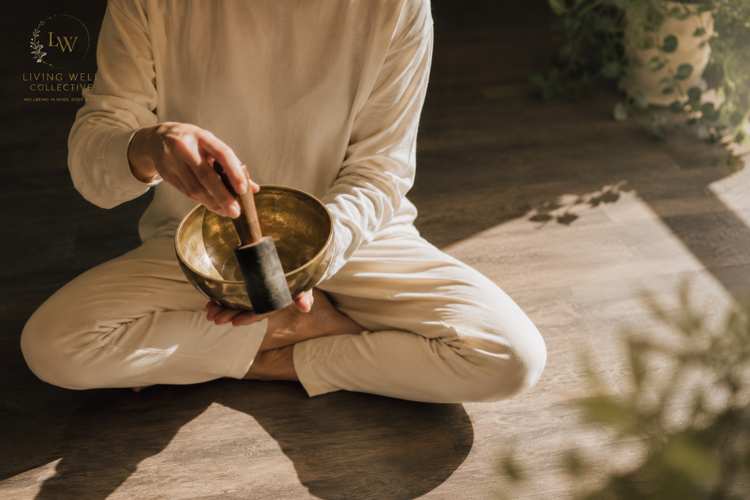 Person sitting cross-legged on a wooden floor, holding a brass singing bowl and a mallet, with natural sunlight and green plants nearby.