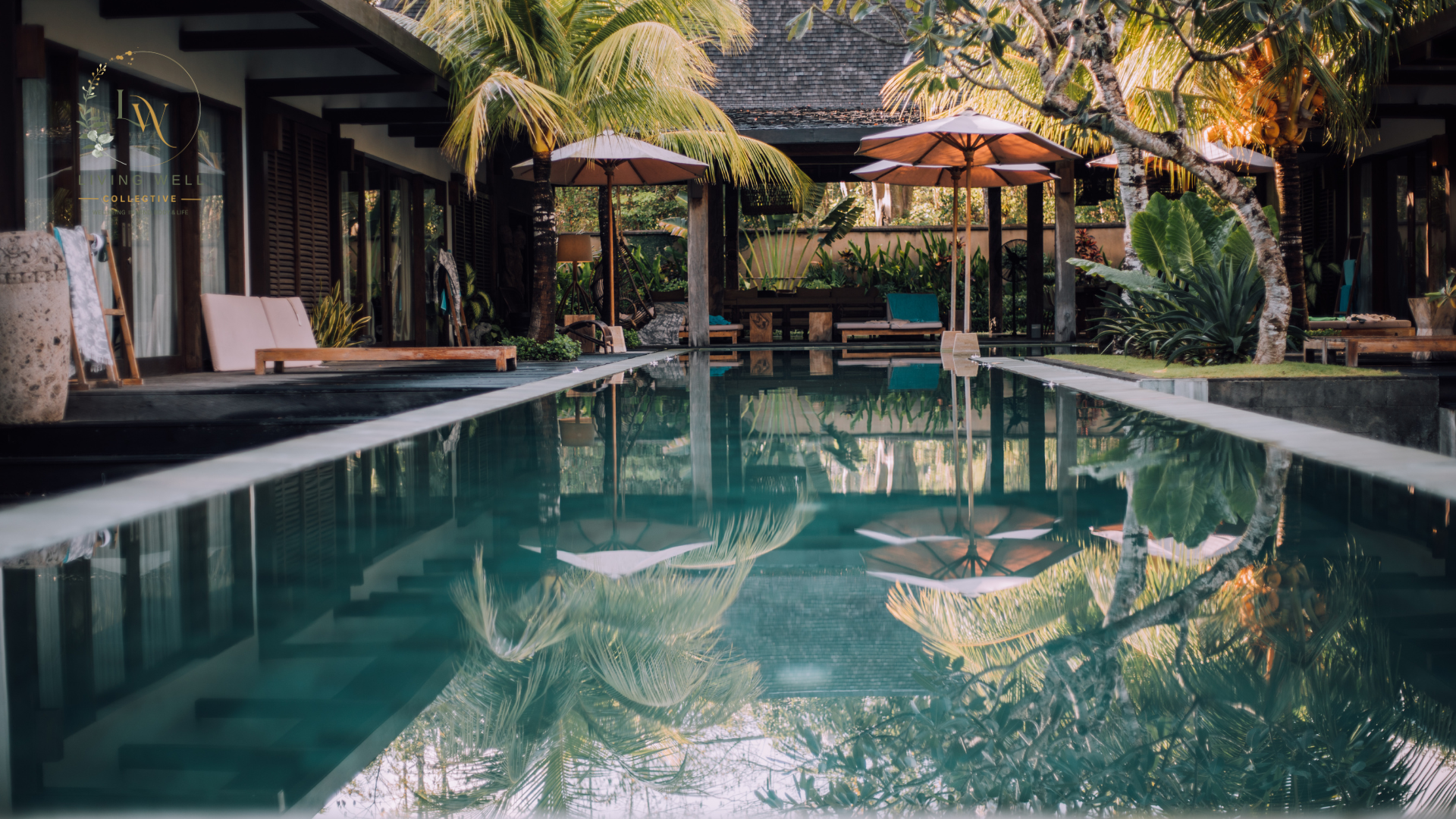 A tropical poolside scene with large umbrellas, palm trees, lush plants, and a reflection of the greenery in the water.